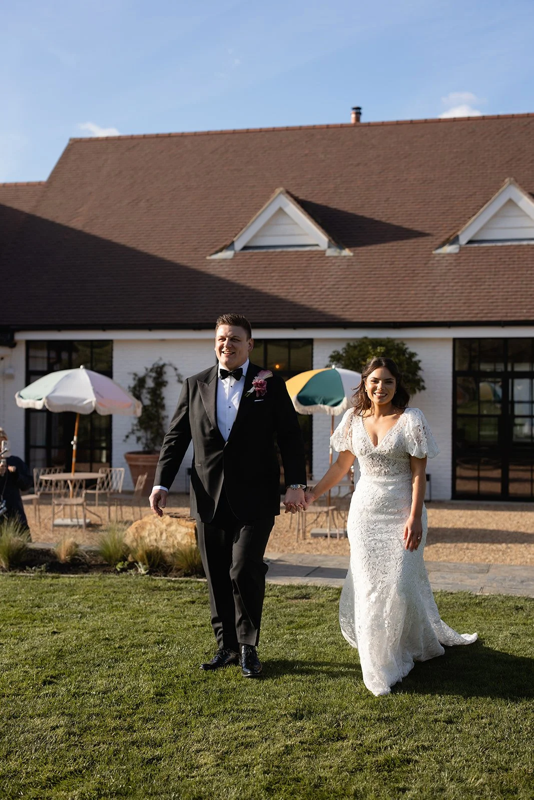 A newlywed couple holding hands and walking outside on a sunny day, with a large house and outdoor seating in the background.