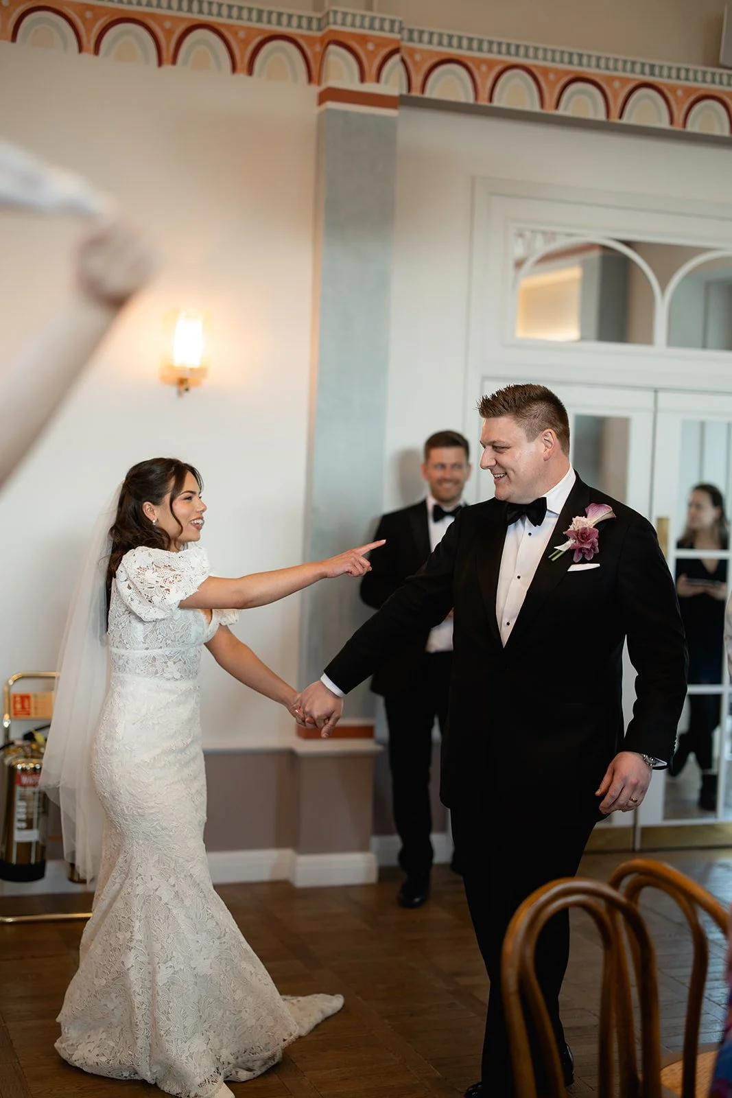 Bride and groom holding hands and smiling during their wedding ceremony, with bridesmaid and groomsman in background, indoors with decorative wall and wooden floor.