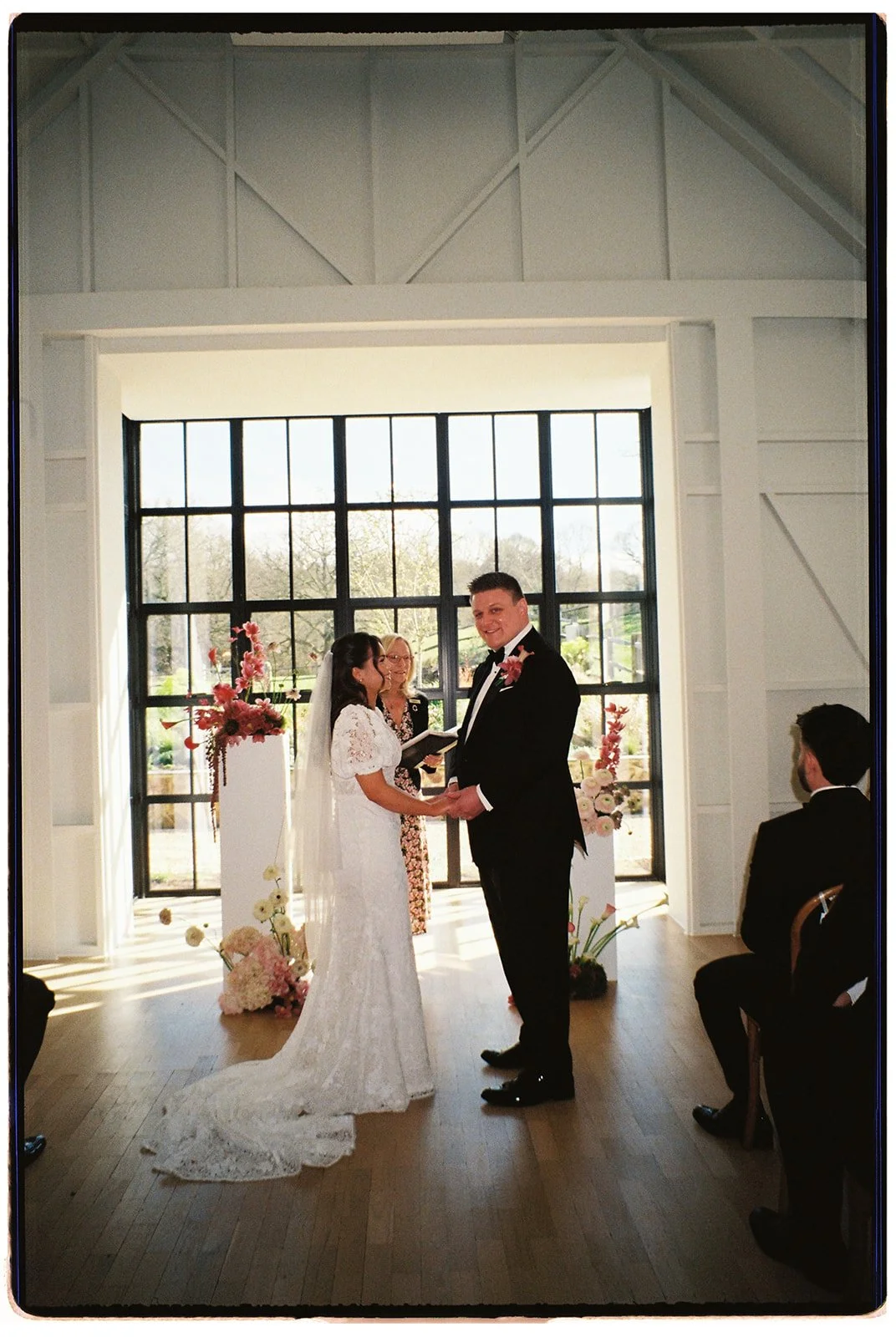 A wedding ceremony taking place indoors with a bride in a white lace gown and a groom in a black tuxedo, holding hands and smiling at each other, in front of large windows with floral arrangements.