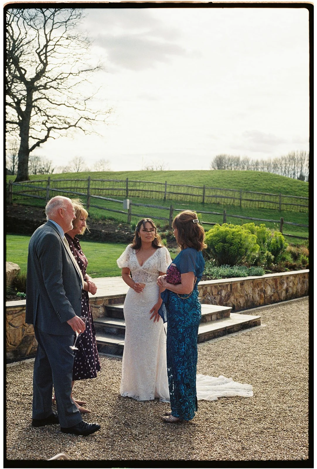 Four people, two men and two women, standing outdoors on a gravel surface, engaged in conversation during a wedding ceremony. One woman is wearing a white wedding dress, and the other has on a blue dress. The men are dressed in suits. The background 