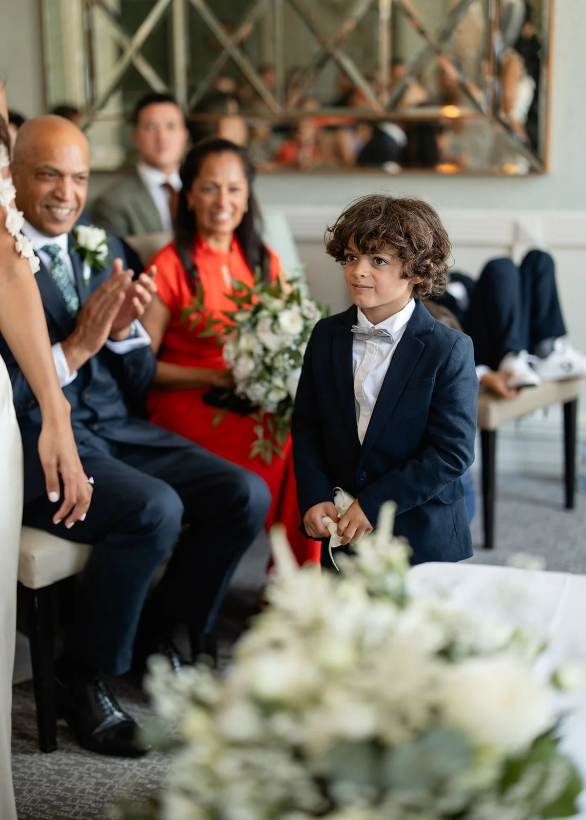 A young boy dressed in a navy blue suit with a white bow tie, standing at a formal event, surrounded by seated adults who are smiling, clapping, and holding a bouquet of flowers.