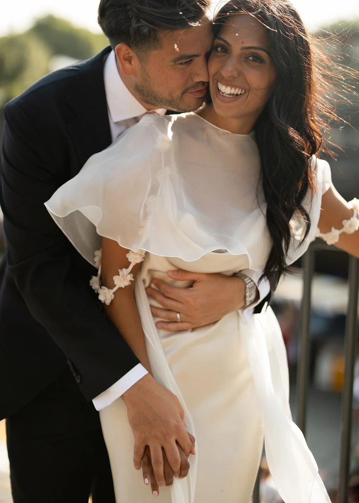 A couple is celebrating their wedding outdoors, with the groom hugging the bride from behind and both smiling happily.