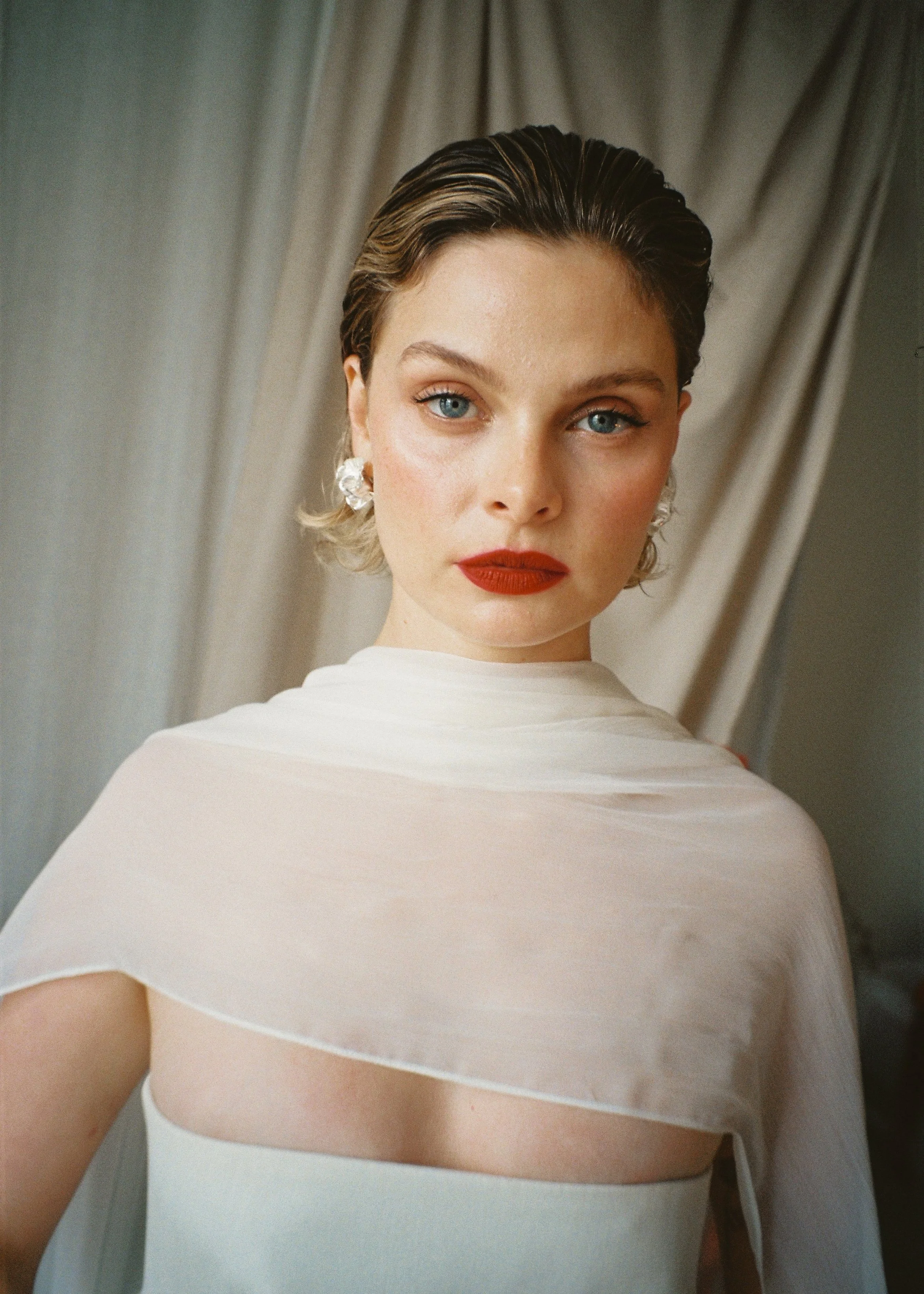 A woman with short, wet-looking hair, blue eyes, and red lipstick wearing a white dress with a sheer cape, standing against a neutral background.