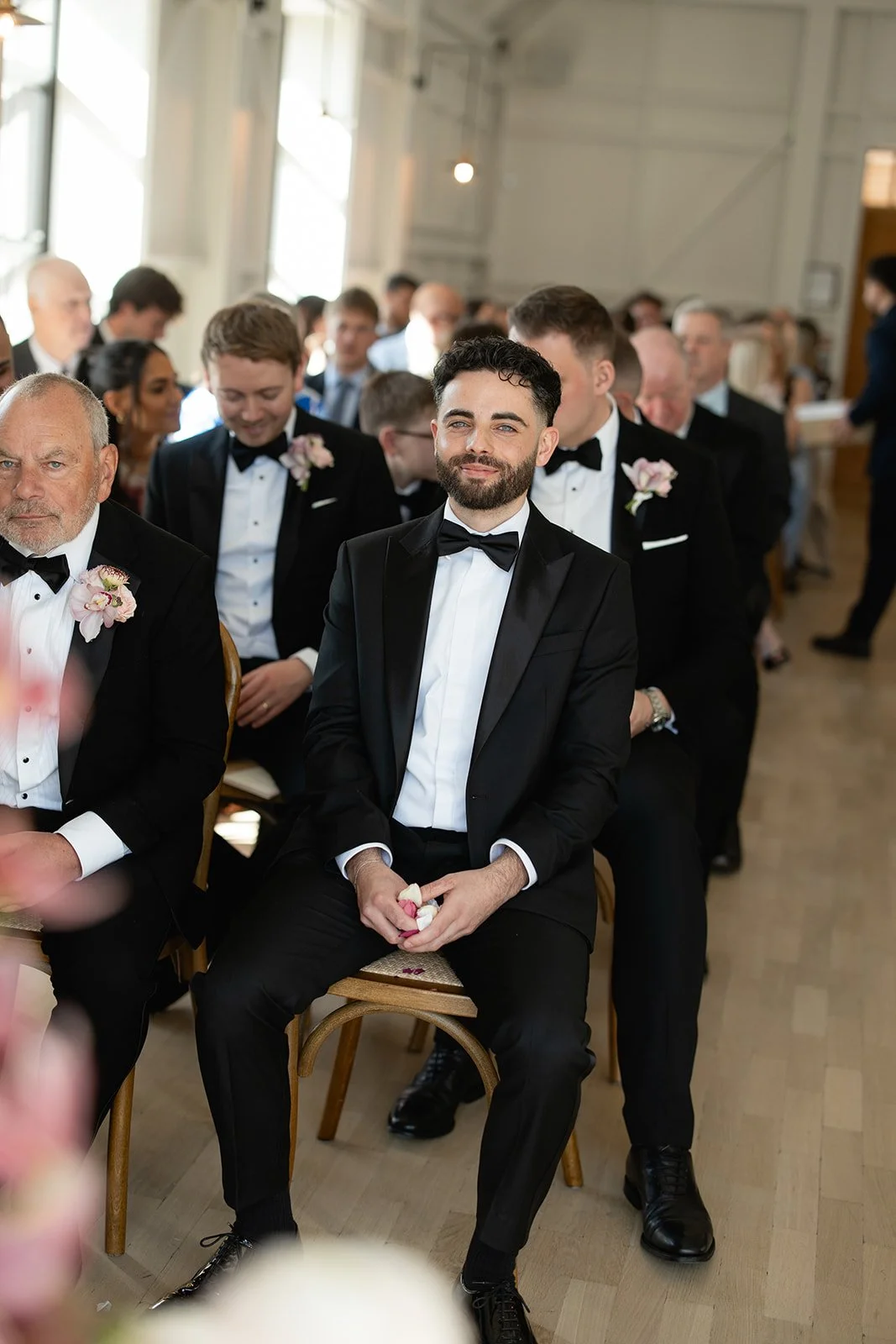 A group of men dressed in formal black tuxedos with bow ties, seated in a brightly lit room during a wedding ceremony. The man in the center is smiling, holding a small gift or item in his hands.
