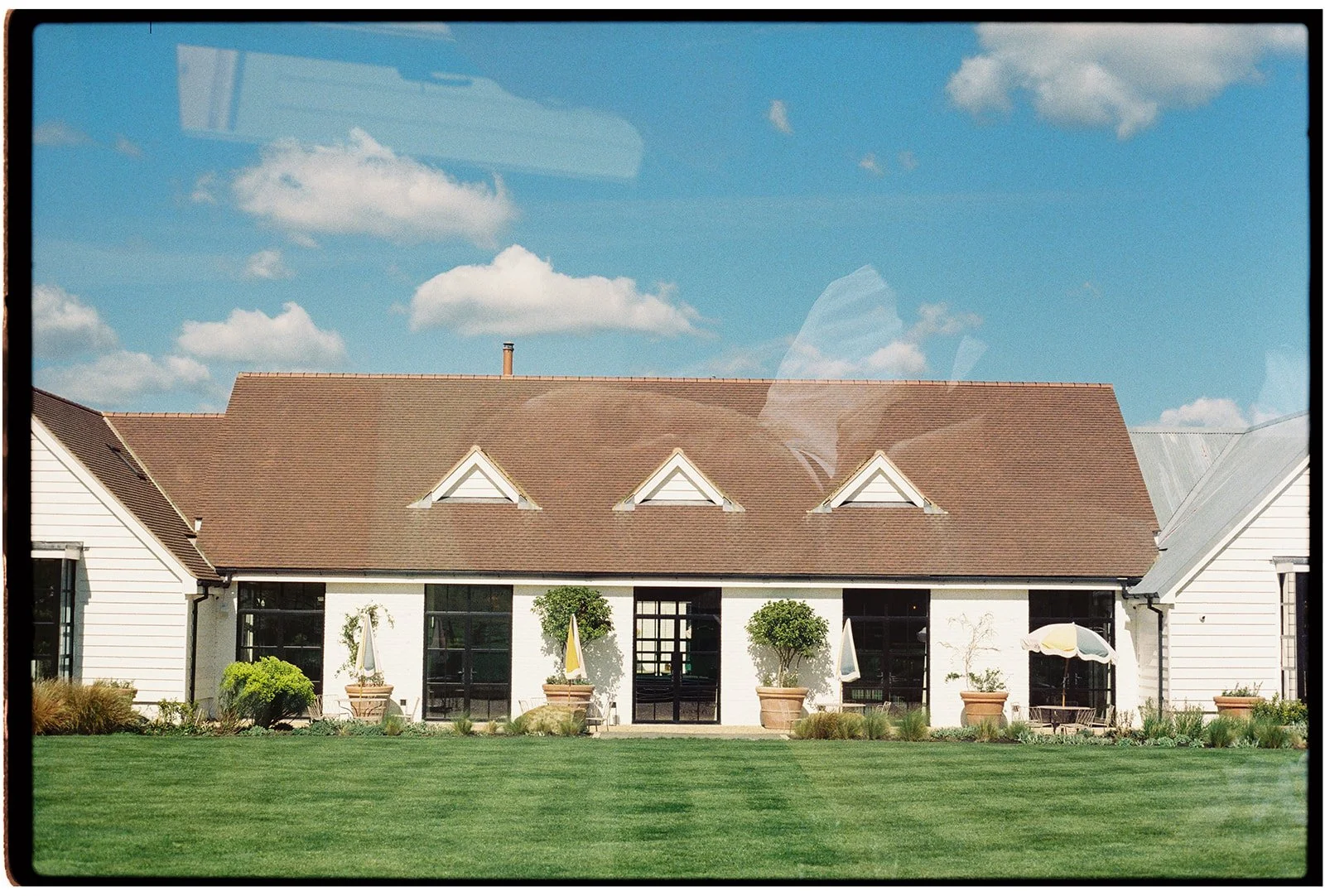 A modern house with white exterior walls, a brown sloped roof with gabled windows, large glass doors, and a well-maintained green lawn in the foreground under a blue sky with some clouds.