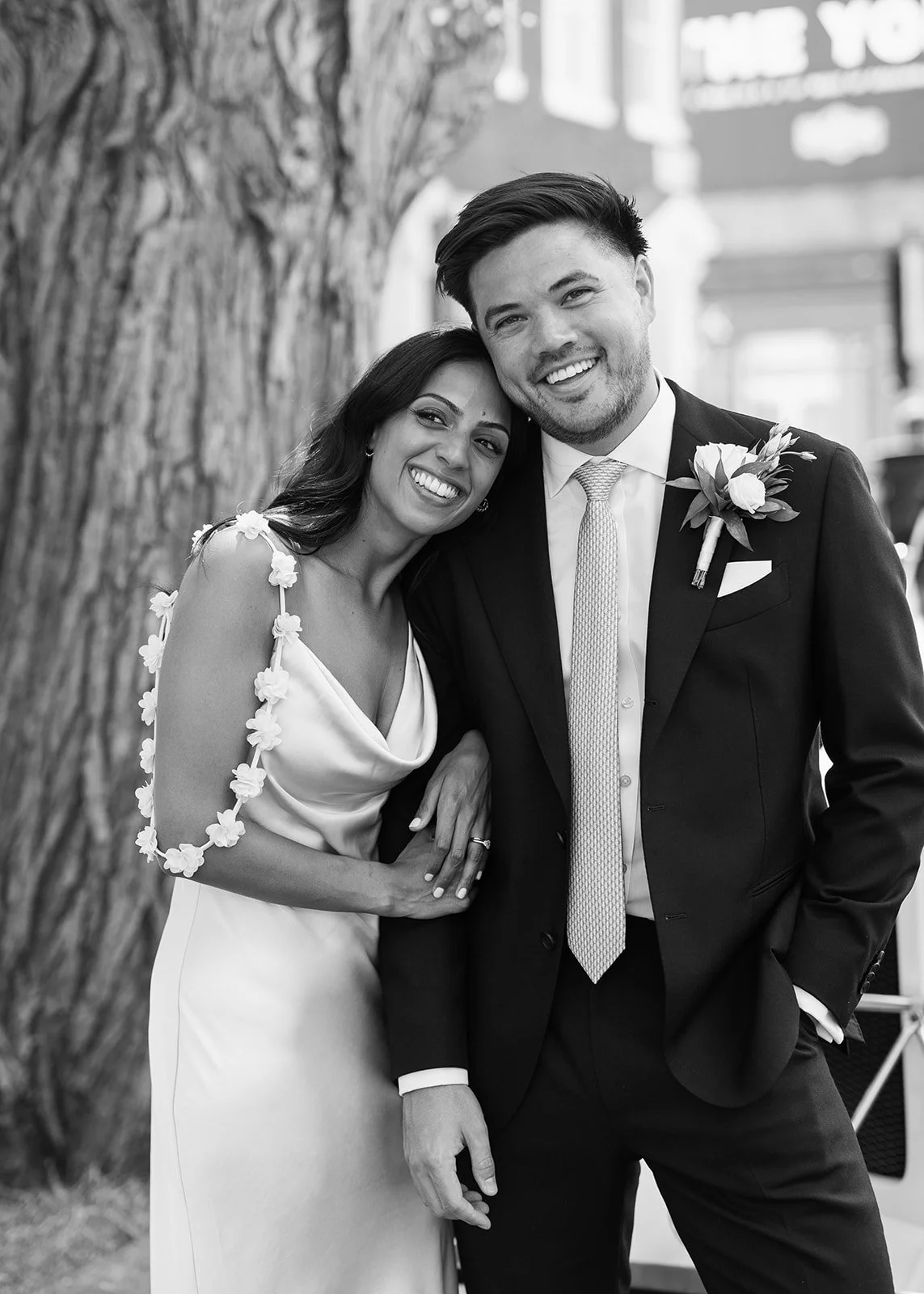A happy couple on their wedding day, smiling and embracing each other outdoors, with a tree and city buildings in the background.