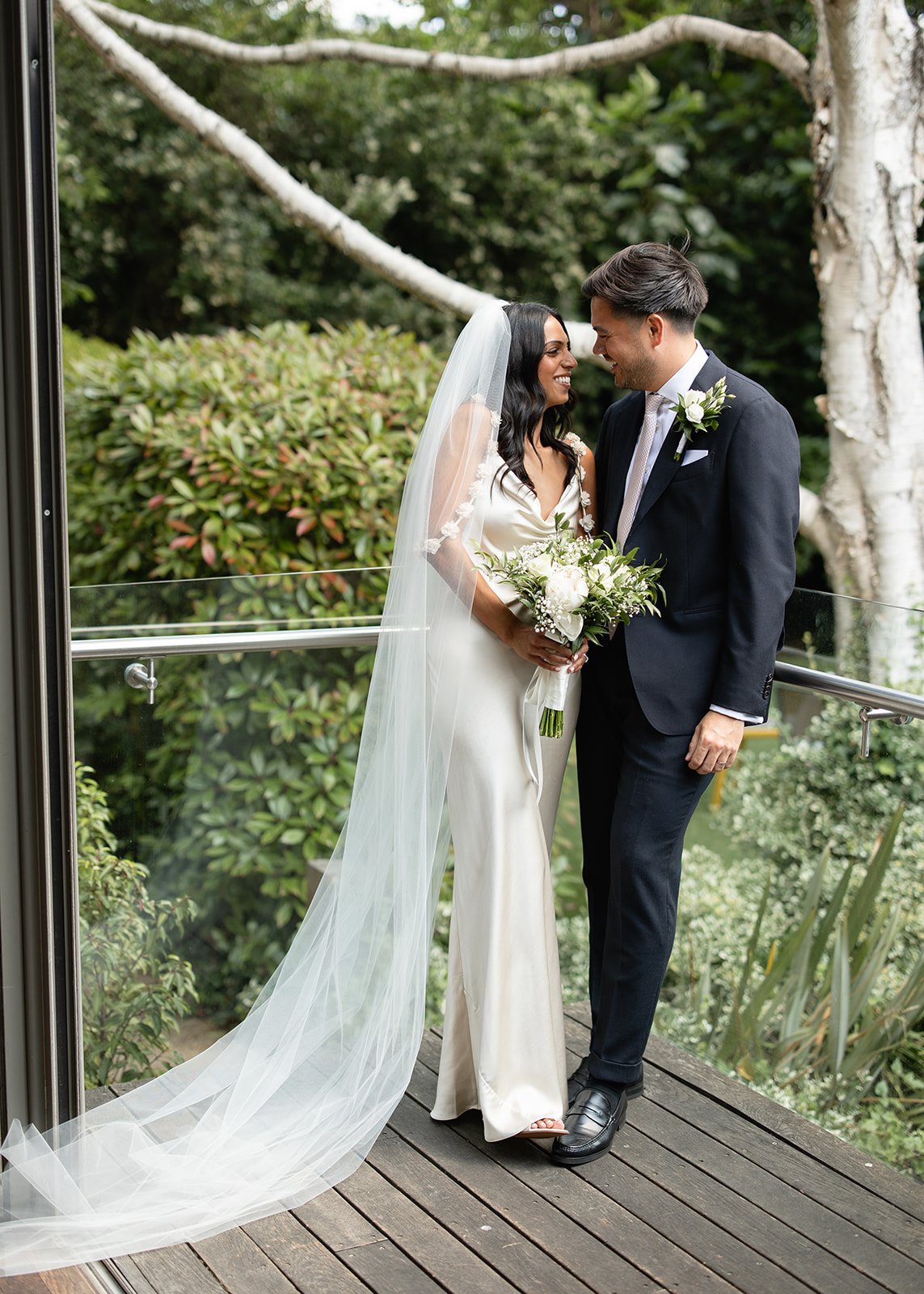 A bride and groom sharing a moment outside on a wooden deck, with a lush green garden in the background. The bride is wearing a white wedding dress and veil, holding a bouquet, and the groom is dressed in a black suit with a white shirt and boutonnie