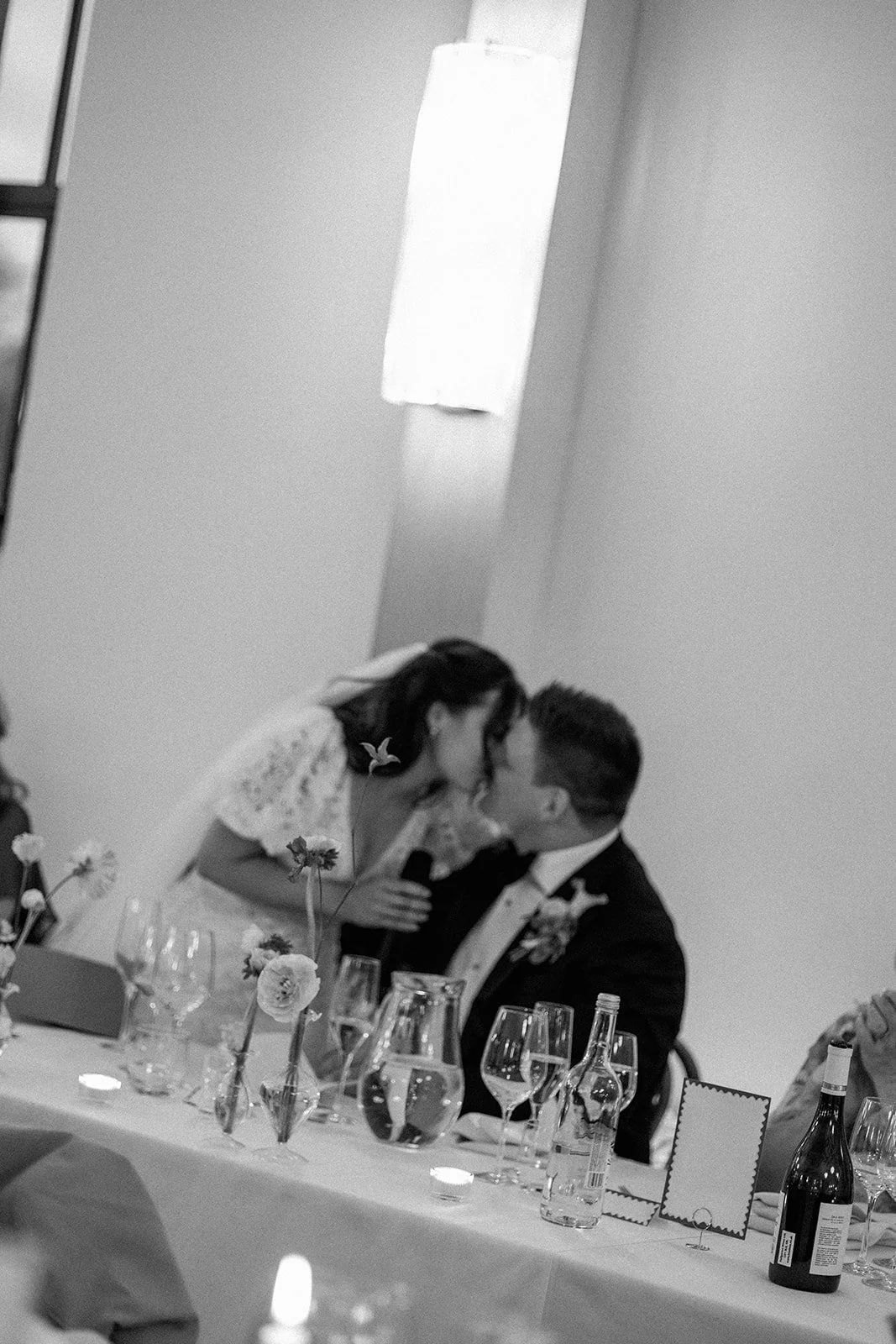A black and white photo of a couple at a wedding reception, leaning in for a kiss. The woman is wearing a lace dress and has flowers in her hair, and the man is in a tuxedo with a boutonniere. They are seated at a table decorated with flowers, glasse