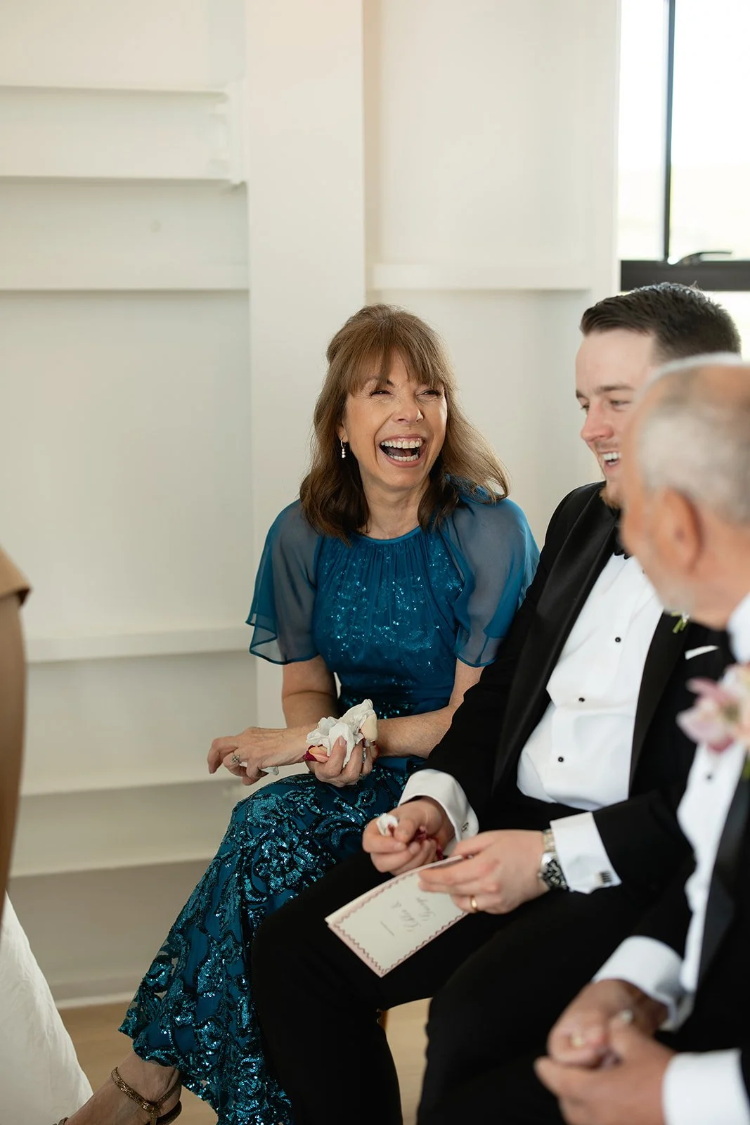 A woman in a blue dress laughing and sitting next to men in formal attire at a wedding or celebration event.