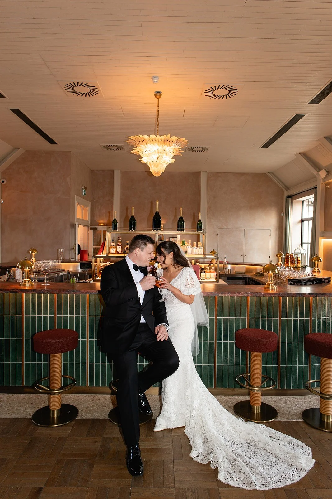 A newlywed couple in wedding attire sharing a toast at a bar, with the bride in a white lace wedding gown and the groom in a black tuxedo, inside a stylish venue with a green-tiled bar and elegant decor.