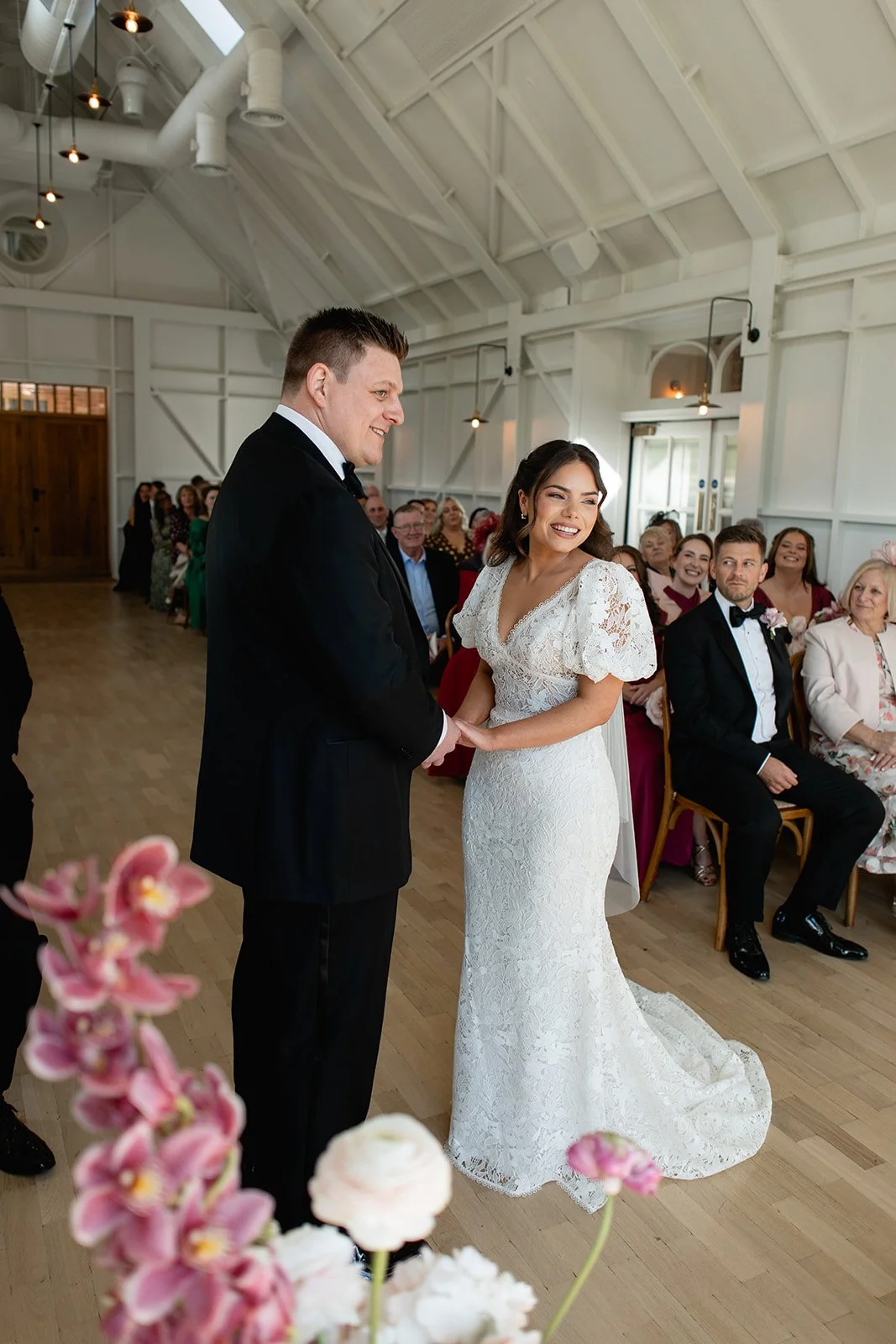A bride and groom hold hands during their wedding ceremony inside a bright, white, wooden venue with guests sitting in chairs behind them. The bride wears a lace white gown, and the groom is in a black tuxedo. Flowers are visible in the foreground.