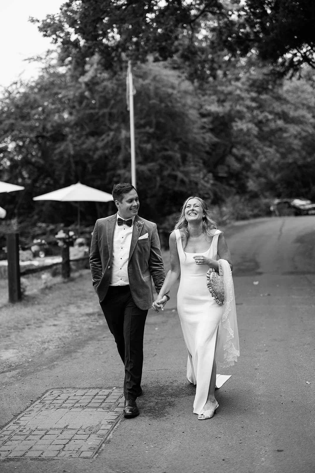 A newlywed couple walking hand in hand outdoors, smiling and looking at each other, with trees and umbrellas in the background.