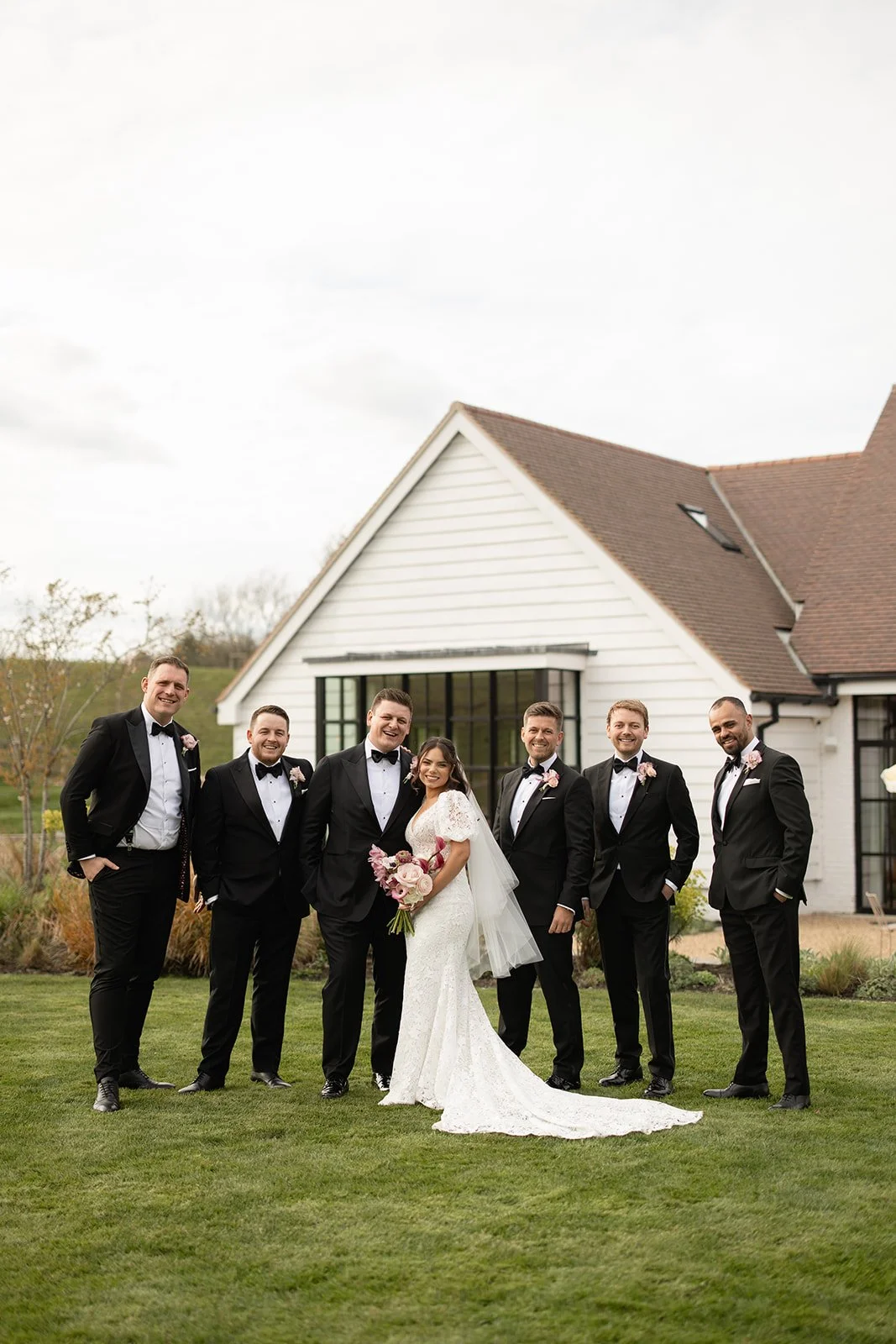 Bride in a white lace wedding dress holding a pink and white bouquet, standing with seven groomsmen in tuxedos outside a white house with a brown roof, green lawn, and cloudy sky.