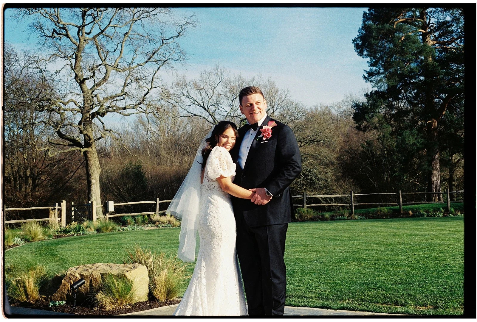 A bride and groom standing together outdoors on a sunny day, holding hands and smiling. The bride is wearing a white wedding gown with lace details and a veil, while the groom is dressed in a black tuxedo with a pink flower boutonniere. There are tre