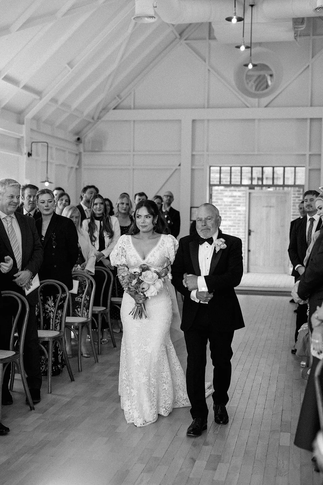A bride in a lace wedding gown holding a bouquet walking down the aisle with her father at a wedding ceremony, surrounded by seated and standing guests in a rustic venue.