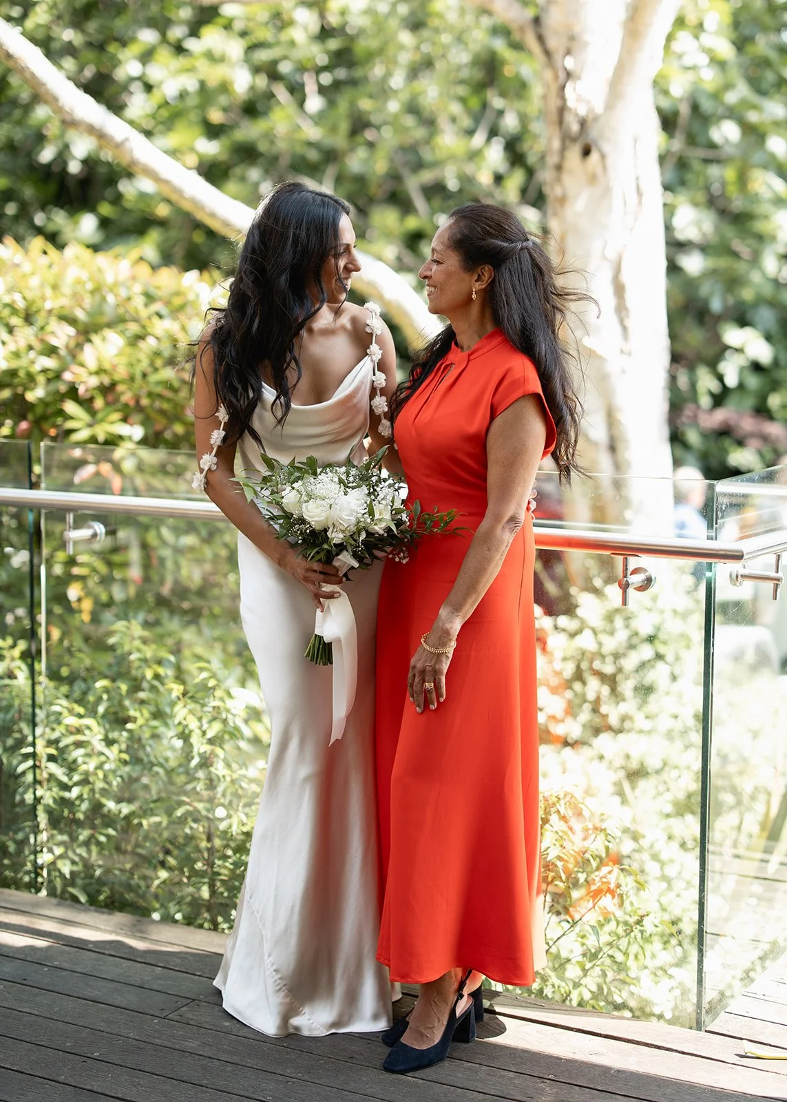 A woman in a white dress holding a bouquet, smiling at a woman in a red dress, outdoors with greenery in the background.