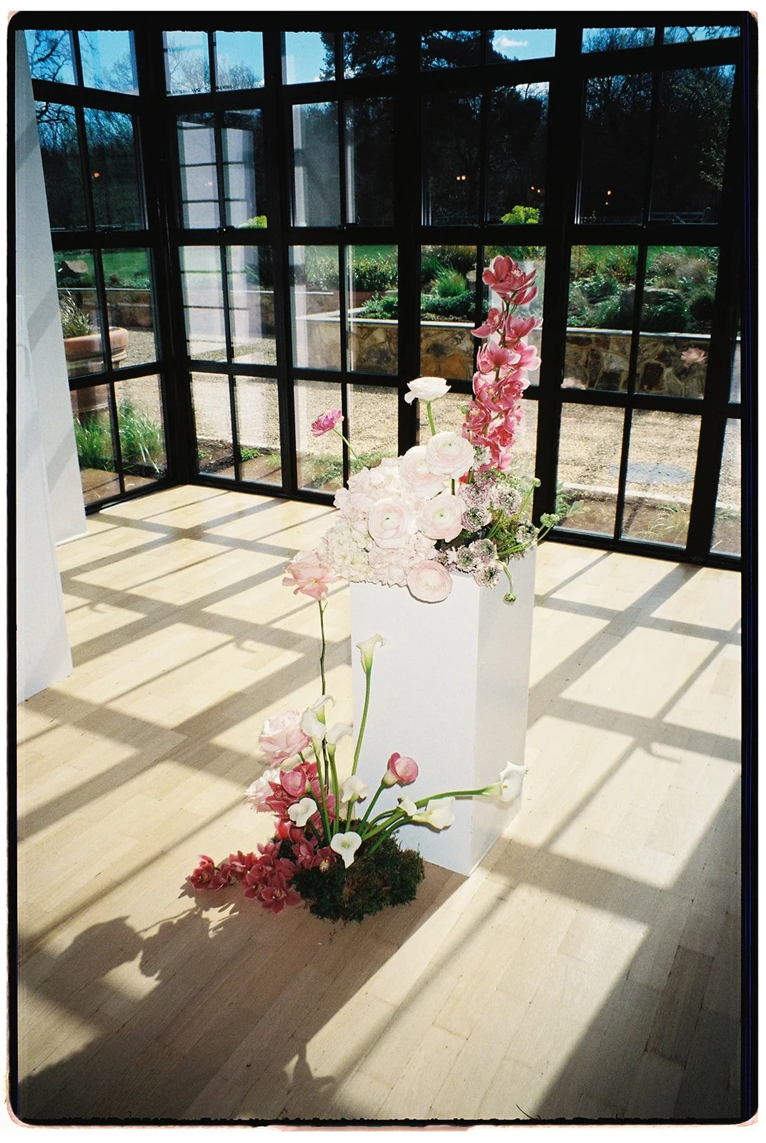 Flower arrangement with pink and white flowers inside a sunlit room with large black-framed windows, overlooking a garden.