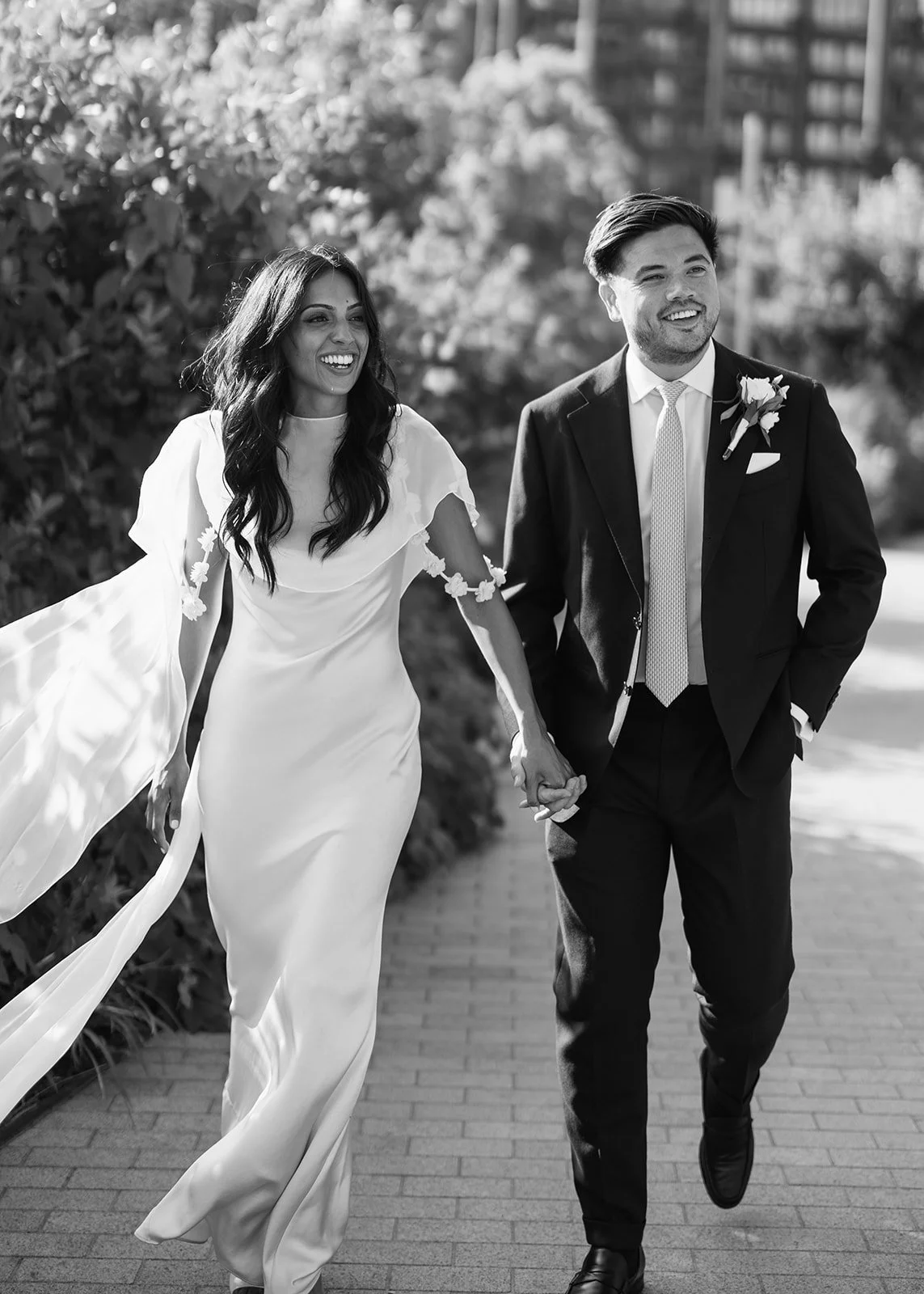 A bride and groom walking hand in hand outdoors, smiling, in a black and white photo. The bride is wearing a flowing wedding dress with floral accents on the sleeves, and the groom is dressed in a suit with a boutonniere.