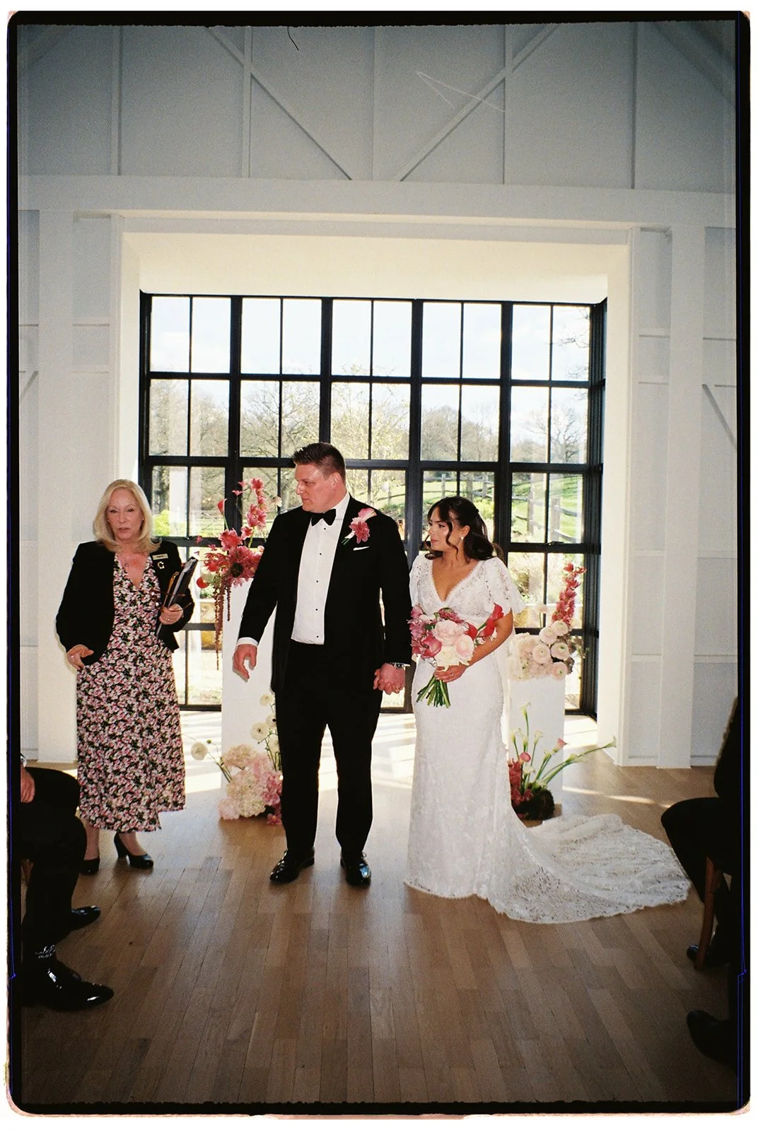 A bride and groom standing in front of a large window during their wedding ceremony, with a woman officiant on the left speaking and floral arrangements behind them.