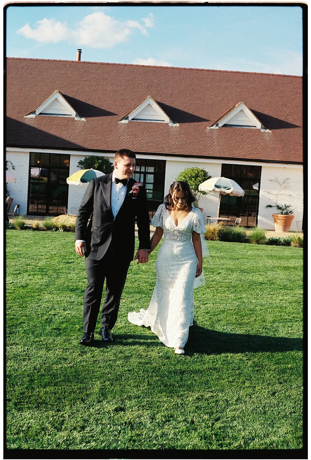 A bride and groom holding hands and walking on a grassy lawn outside, with a building and umbrellas in the background, during a wedding celebration.