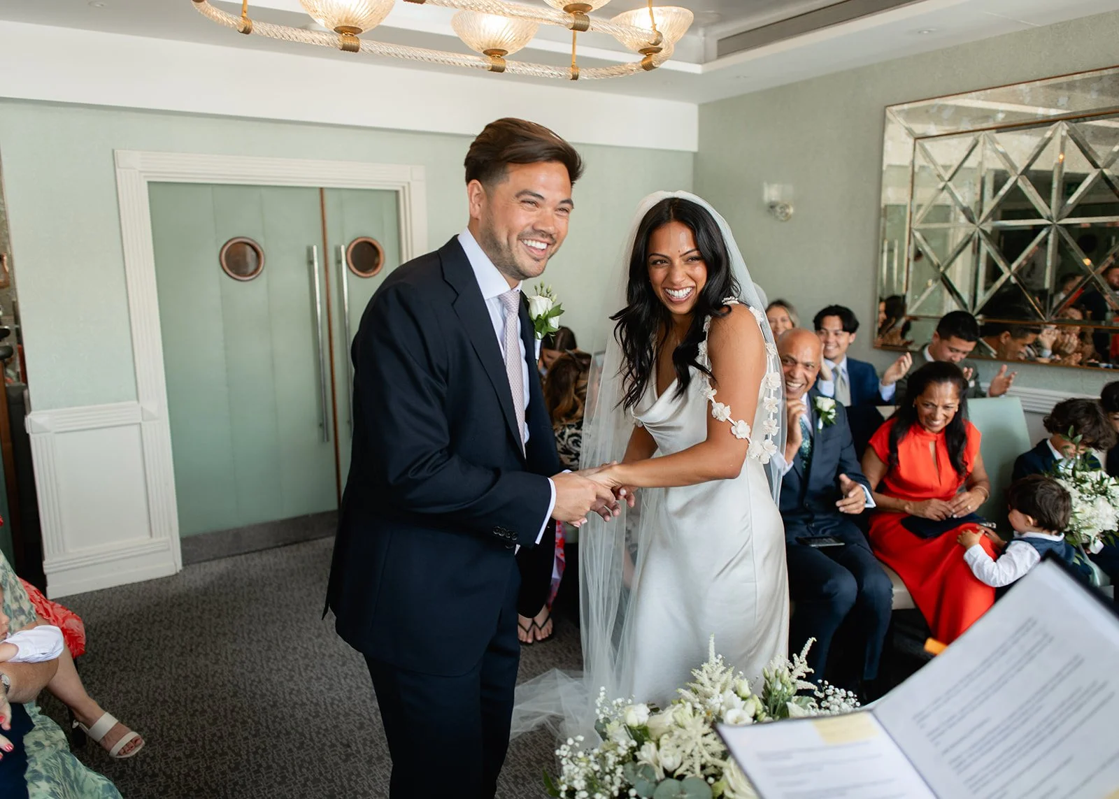 A bride and groom smiling and holding hands during their wedding ceremony in an indoor venue surrounded by seated guests.