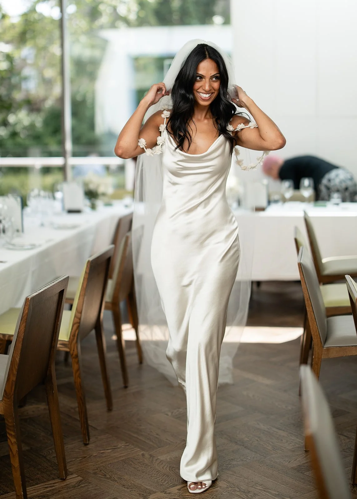 A woman in a satin dress and veil walking in a dining room with tables and chairs, smiling.
