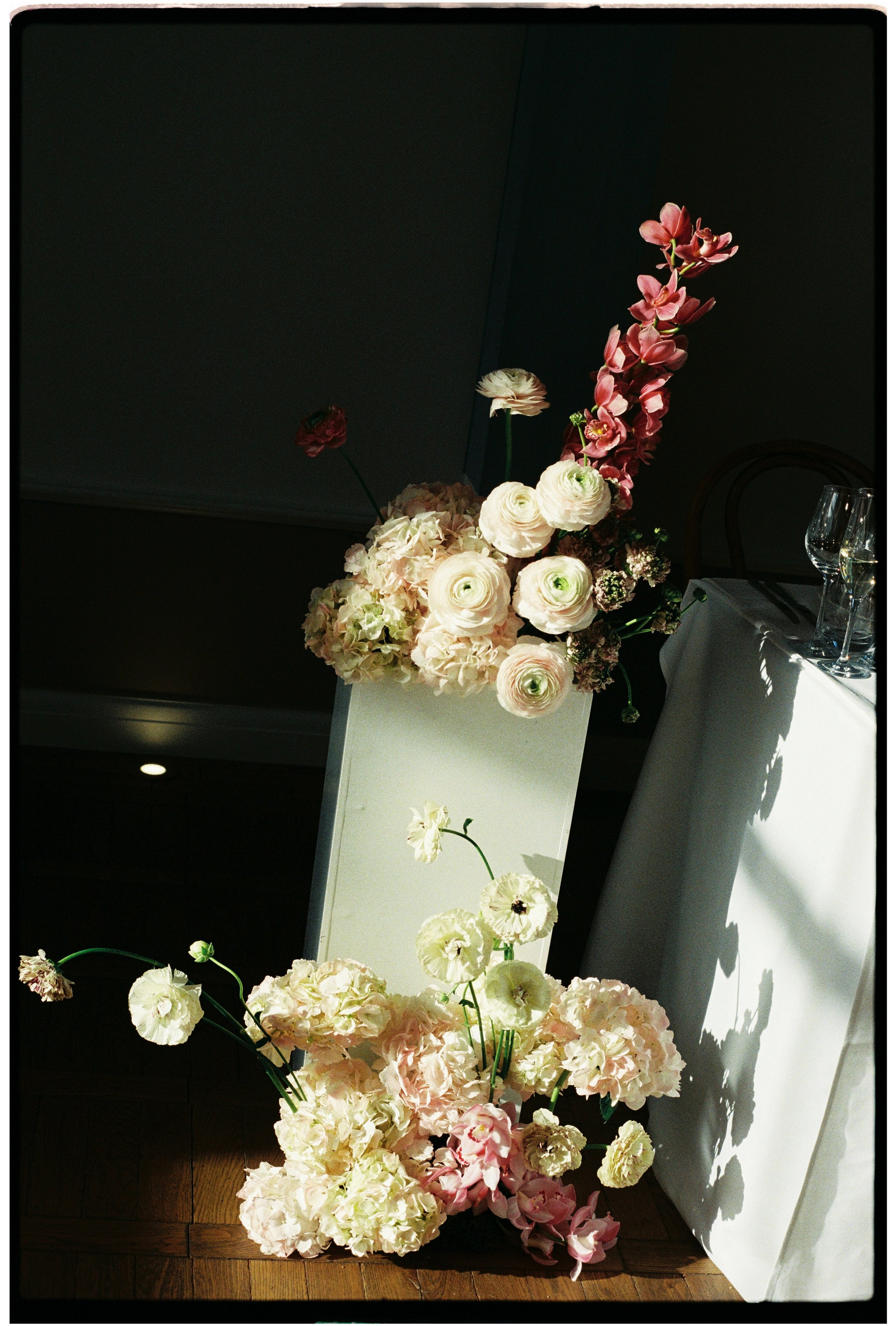 Arrangement of pink, white, and cream flowers, including hydrangeas, ranunculus, and orchids, placed on a white stand next to a table with champagne flutes.