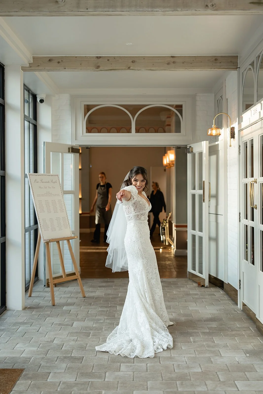 A bride in a white lace wedding dress and veil smiling and pointing towards the camera inside a bright wedding venue with large windows, a sign on a wooden easel, and staff in the background.
