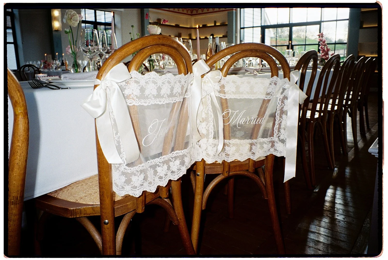Wedding reception table set with glassware and floral decorations, with a chair in the foreground draped with a lace and ribbon sash reading 'Just Married' in elegant script.