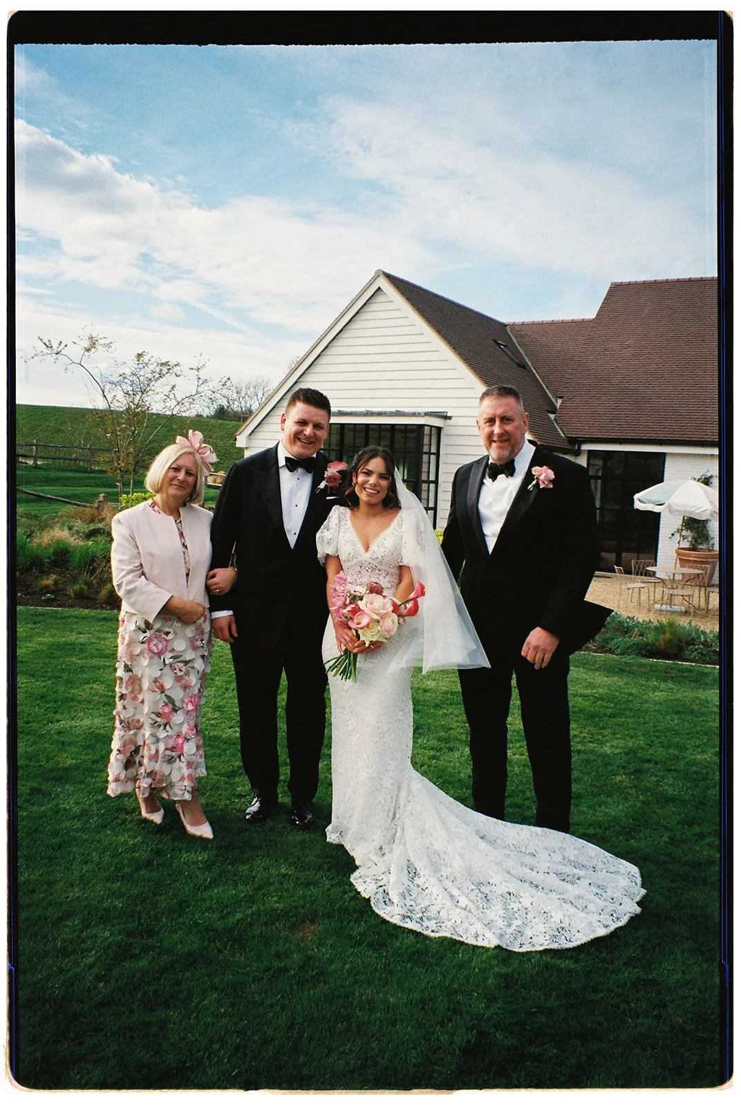 A bride and groom in wedding attire standing outside with two other people, a woman and a man, near a white house with a brown roof and outdoor seating, under a partly cloudy sky.