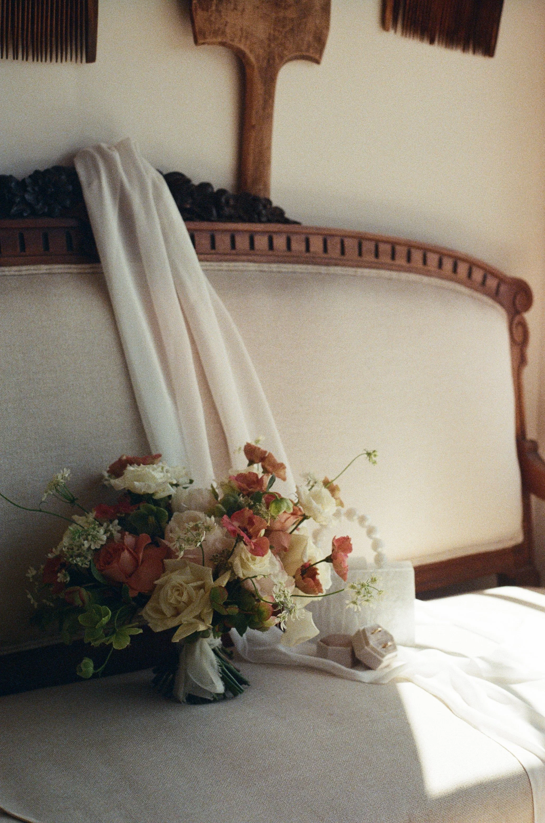 A bouquet of white and pink flowers with greenery, a pearl necklace, and a small jewelry box resting on a white cloth on a vintage bed with a beige headboard in soft natural light.