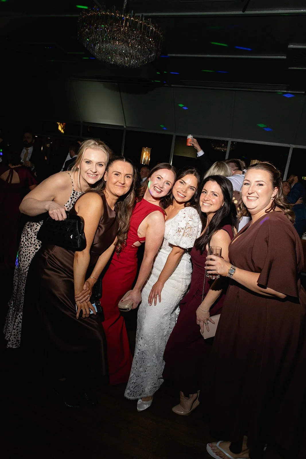 Group of six women in elegant dresses smiling at a party with a chandelier overhead and other guests in the background.