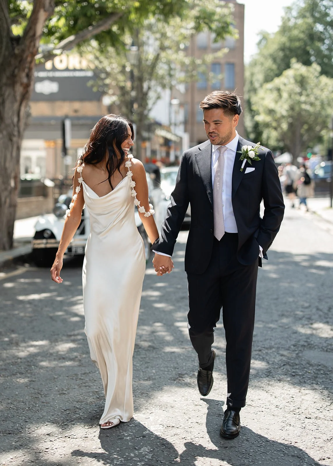 Couple dressed in wedding attire walking hand in hand on a city street, smiling, with trees and buildings in the background.