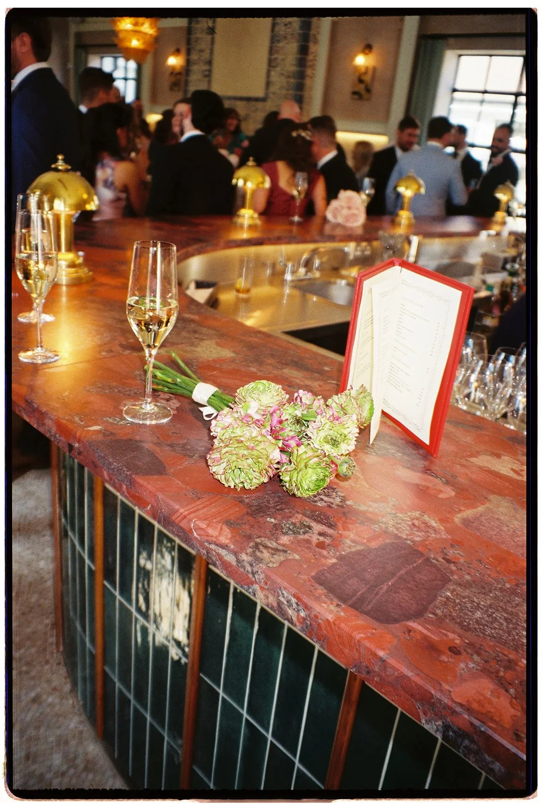 A bar counter with two glasses of champagne, a bouquet of green and pink flowers, and a menu in a red cover, with a crowd of people dressed formally in the background at a social event or celebration.