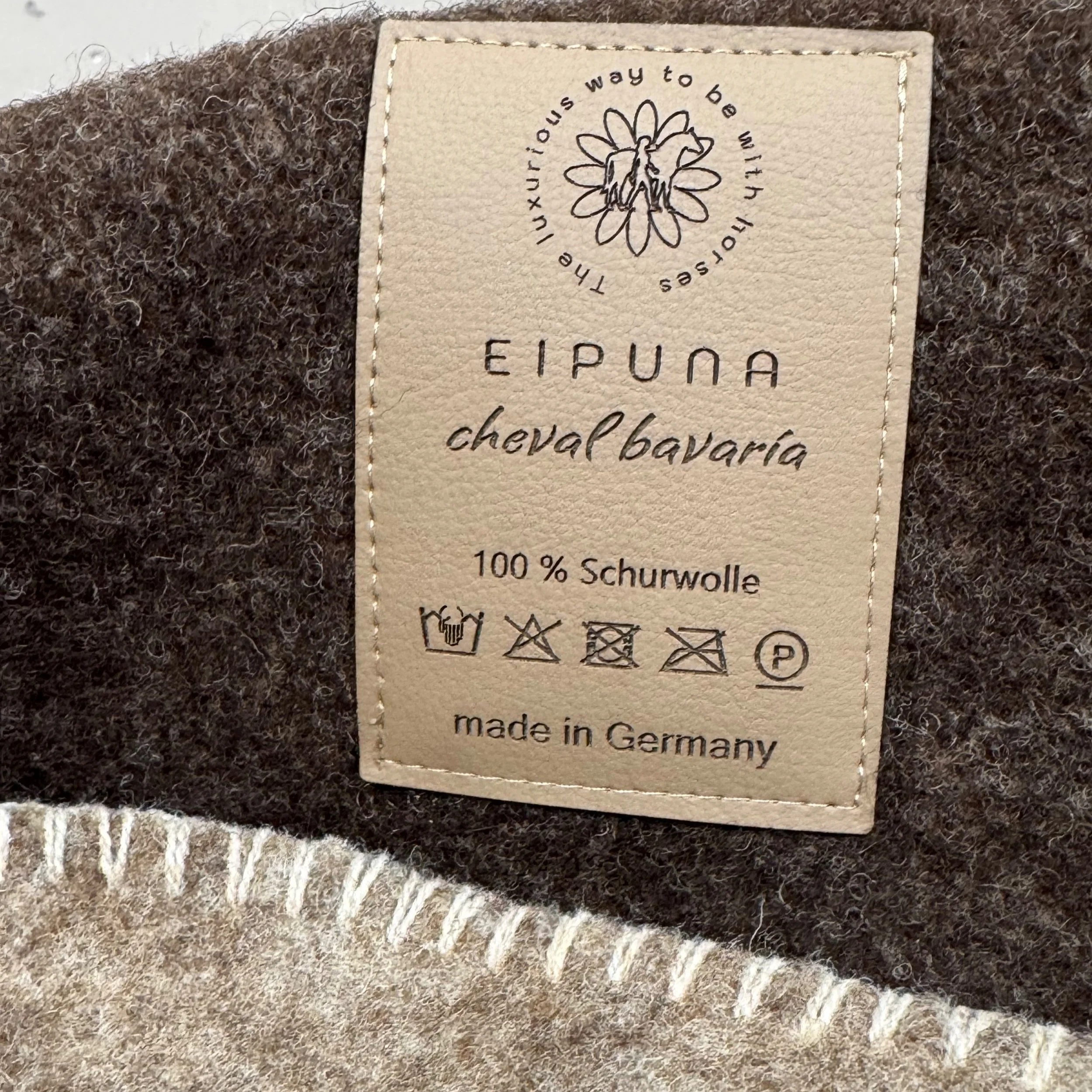 Waschhinweisetikett auf einem braunen Wolltextil, mit Text in Deutsch und Symbolen für die Pflegeanleitung, inklusive der Angabe, dass das Produkt 100 % Schurwolle ist, hergestellt in Deutschland.