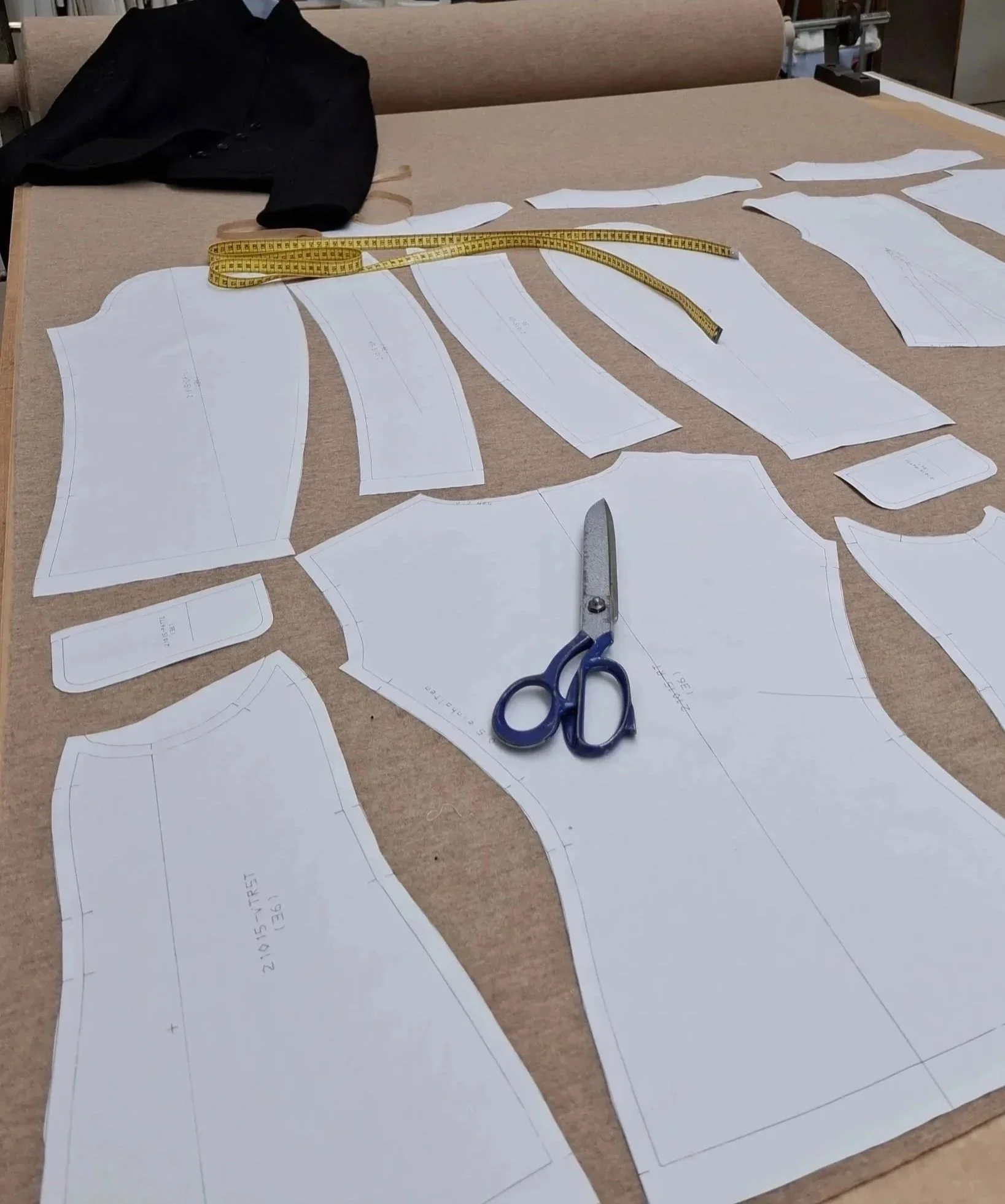 Schnittmusterpapiere für eine Kleidung auf einem Arbeitstisch, mit Schere, Maßband, schwarzem Kleidungsstück und Schneidetisch im Hintergrund.