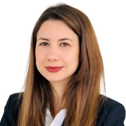 A woman with long brown hair wearing a black blazer and white shirt, smiling at the camera against a white background.
