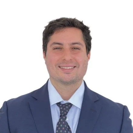 A man with dark hair wearing a navy suit, light blue shirt, and a patterned tie, smiling against a white background.