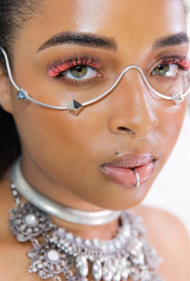 Close-up of a woman wearing colorful makeup, unique jewelry including a nose ring, a choker, and layered necklaces, with a distinctive metal eyewear that features pyramid-shaped studs.