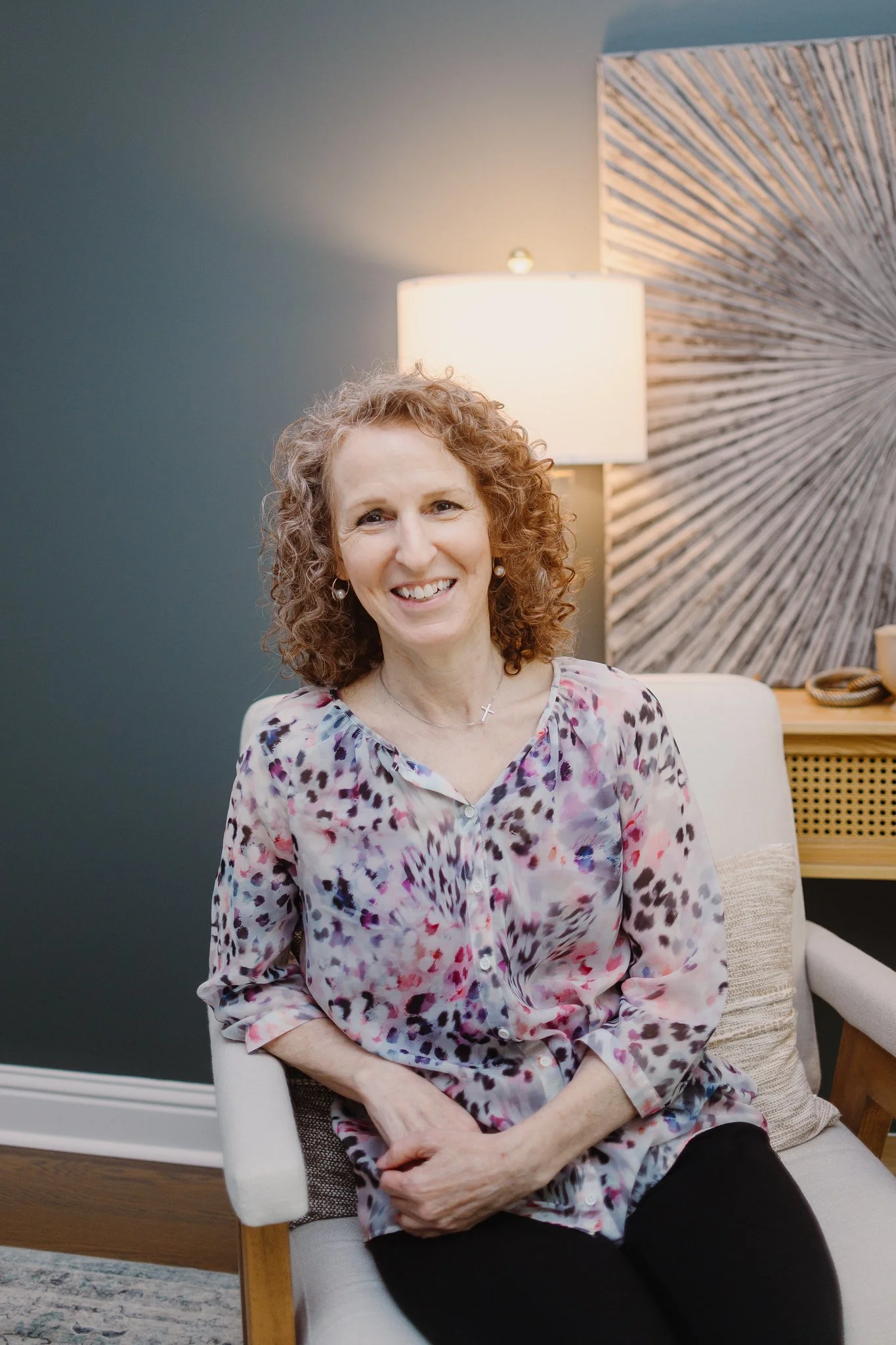 Woman with curly hair sitting on a white armchair in a cozy living room, smiling at the camera, with a large green plant and window in the background.