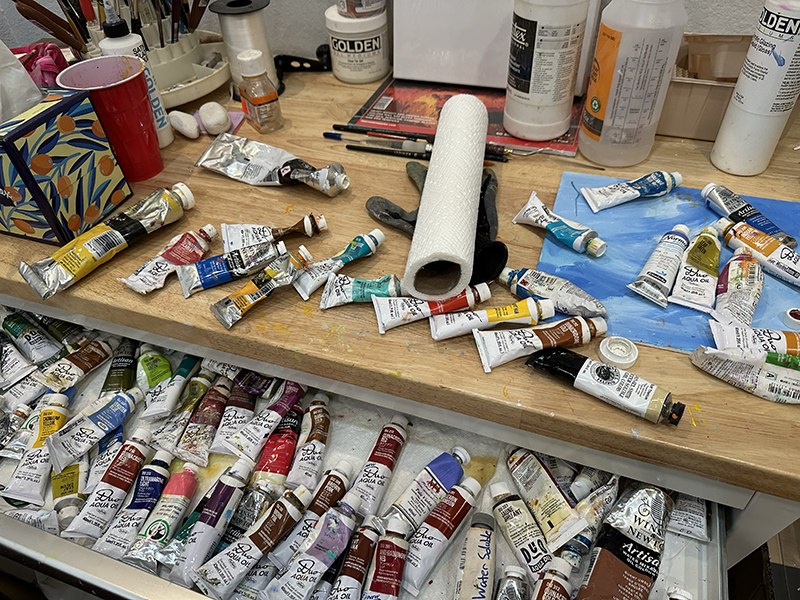 A cluttered art or craft workspace with open paint tube boxes, paint tubes, a paper towel roll, brushes, and various containers and supplies on the wooden surface.