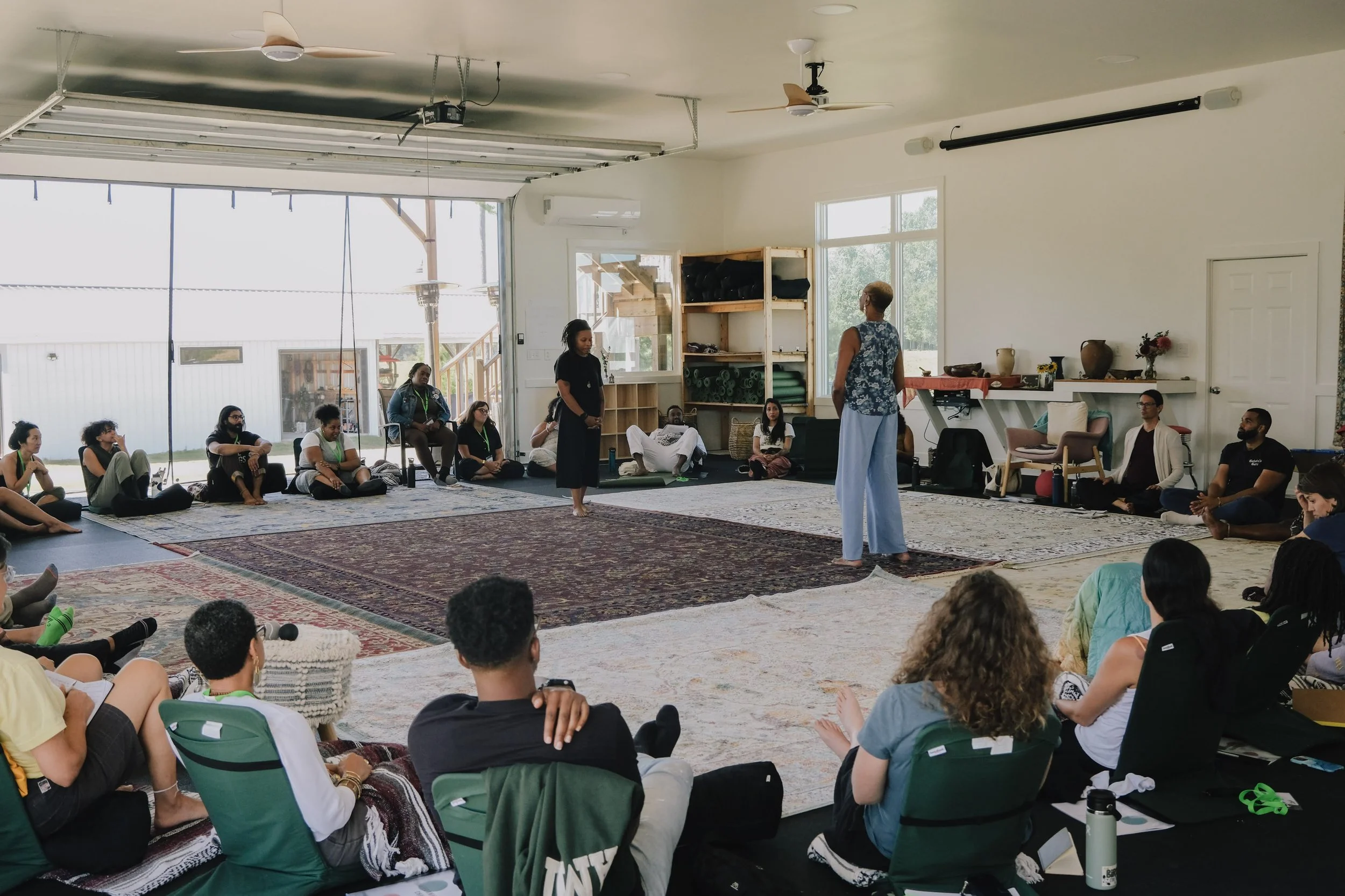 A group of people sitting in a circle on the floor in a bright room with large windows, listening to a woman standing and speaking in the center. The room has rugs, shelves, and various decorative items.