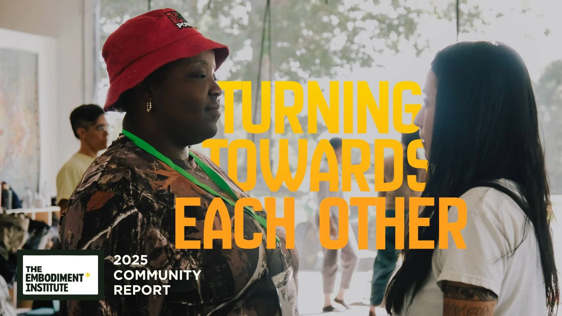 Two women are facing each other and talking at an indoor event. One woman has dark skin, is wearing a red hat, patterned clothing, earrings, and a green lanyard. The other woman has light skin and long black hair. The background shows windows with trees outside, and a person in a yellow shirt is visible in the background. Overlaid text reads 'Turning Towards Each Other.'