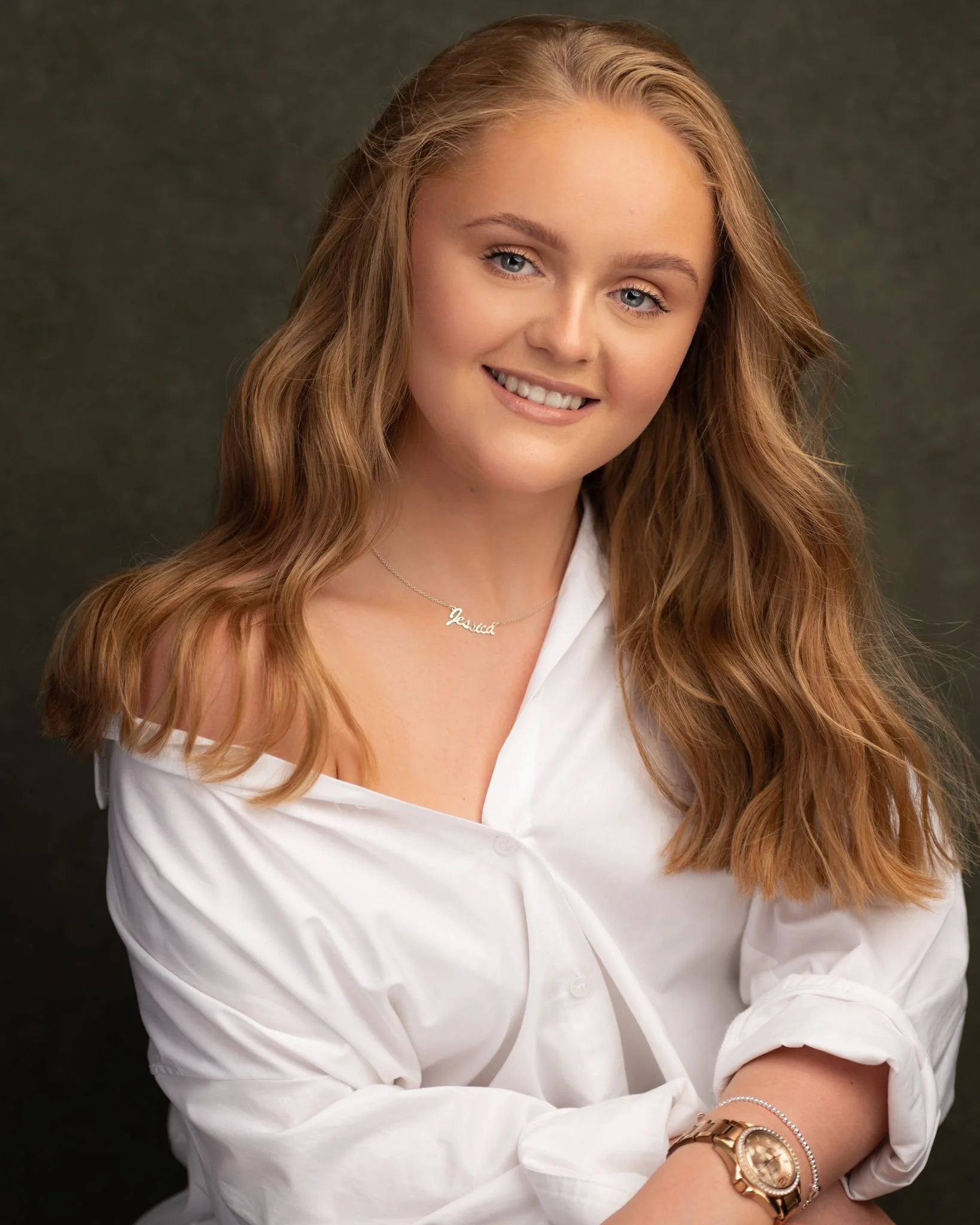 Person wearing a white shirt, smiling, with long wavy hair, and accessorized with a necklace and watch.