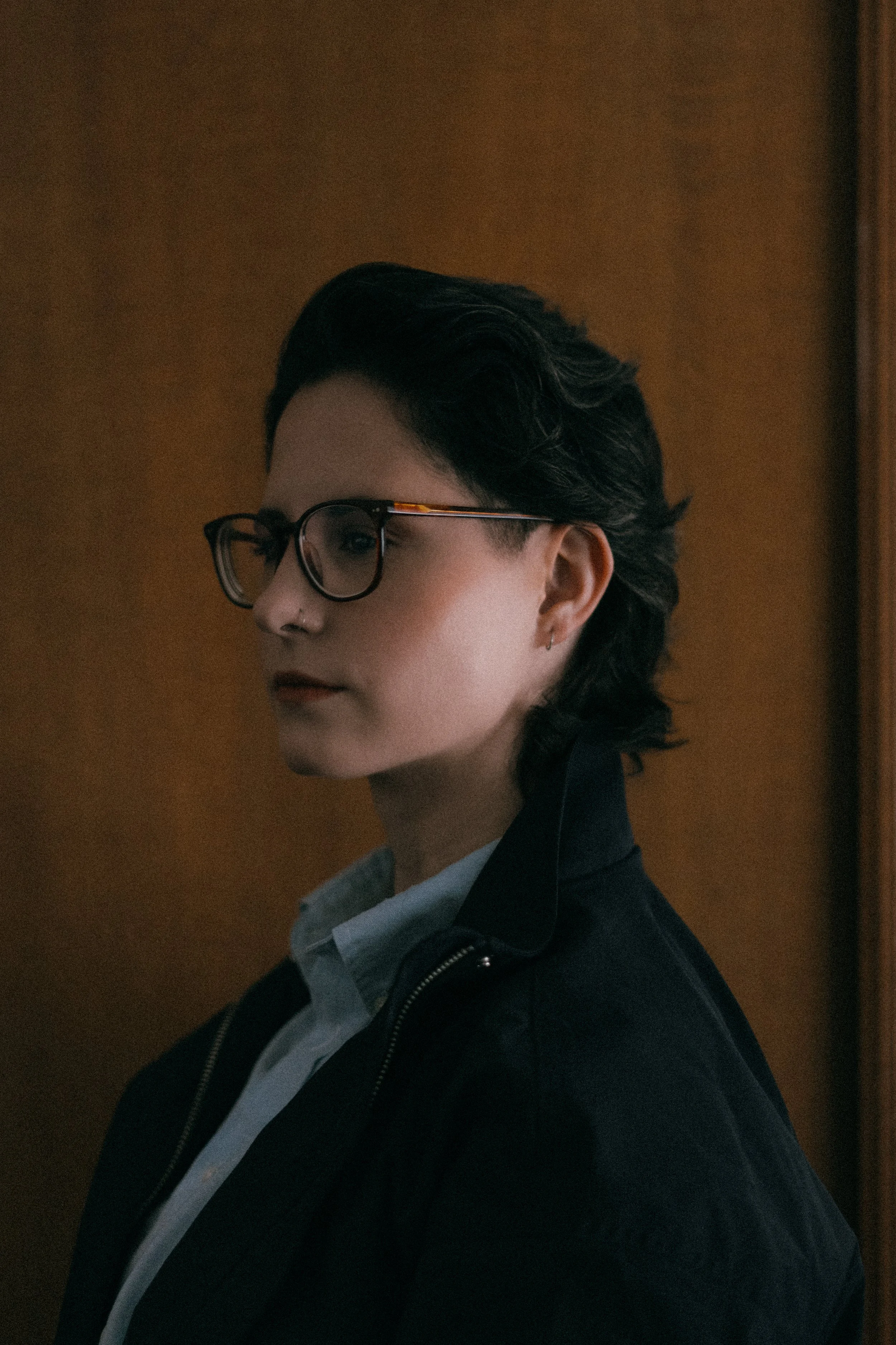 Side profile of a woman (Erin Sandell) with short dark hair, wearing large round glasses, a nose piercing, earrings, a gray hoodie, and a black leather jacket against a brown background.