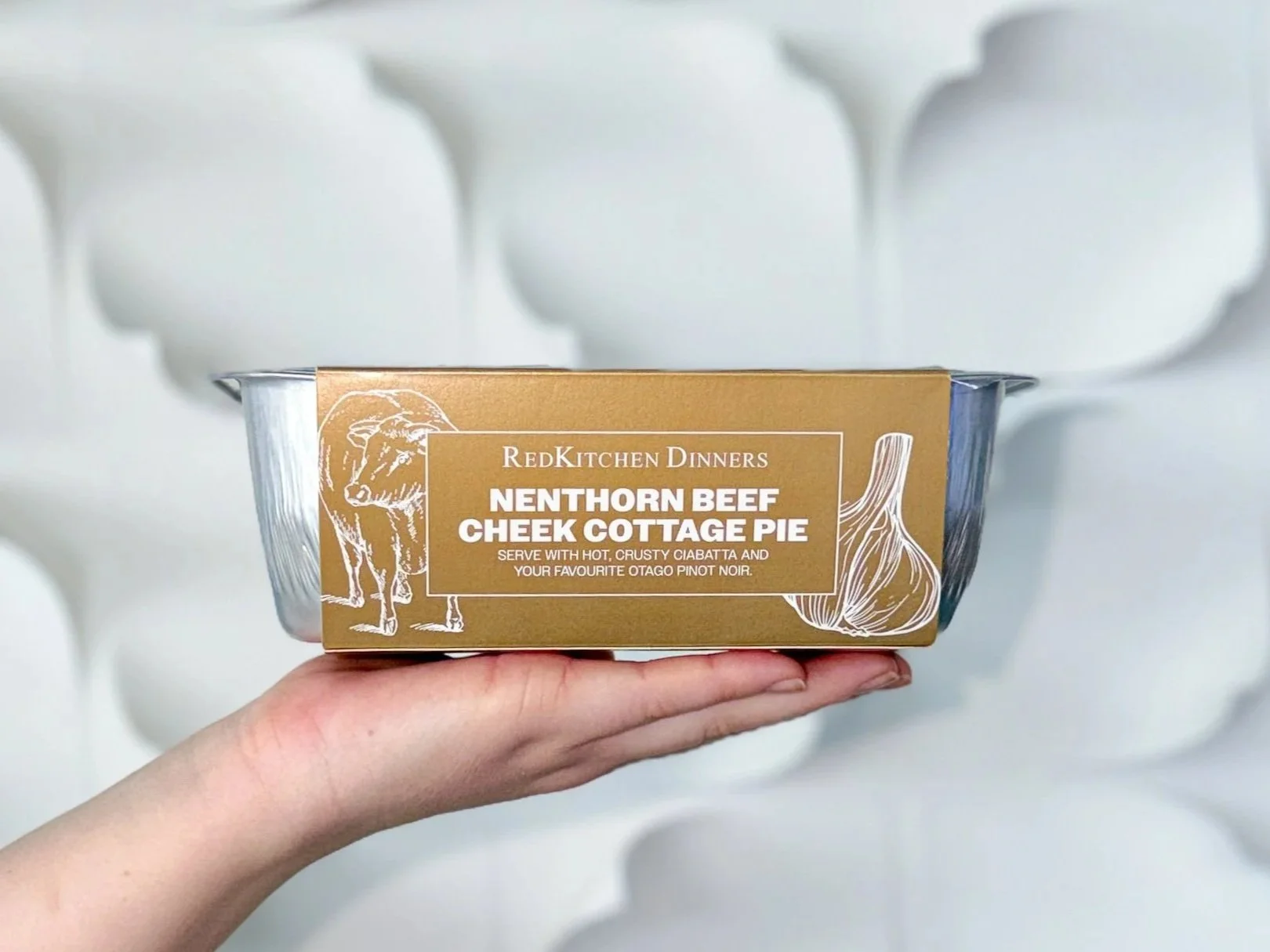 Up close photo of a hand holding a frozen meal of RedKitchen Nenthorn Beef Cheek Cottage Pie in its silver foil container with gold and white packaging.