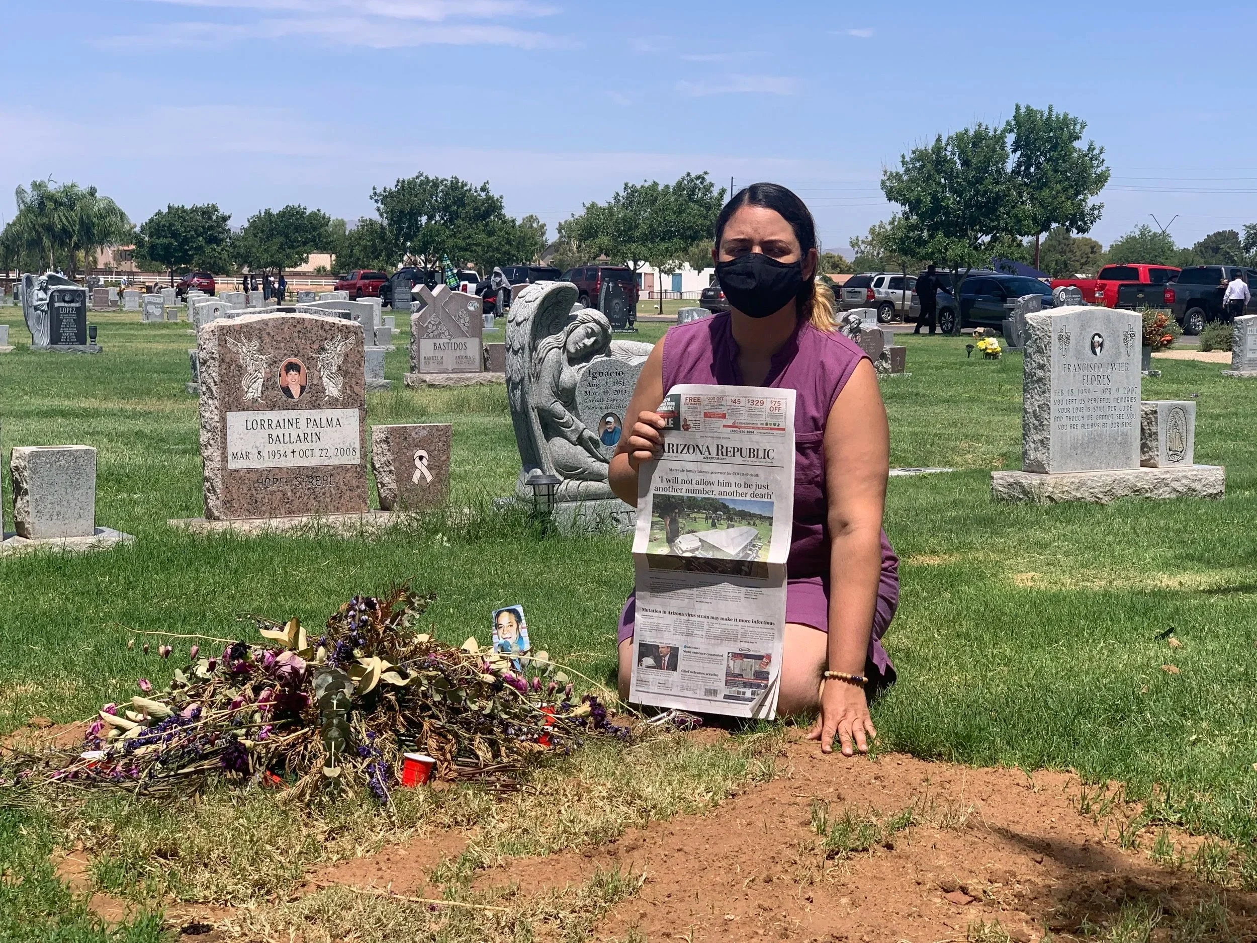 Kristin Urquiza kneeling at her father Mark's grave in a cemetery, wearing a black mask and holding up the front page of the Arizona Republic newspaper featuring the story of his funeral and honest obituary
