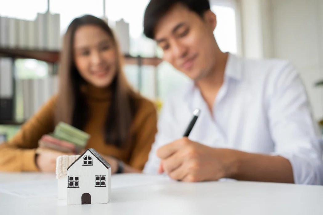Couple signing real estate contract with house model in foreground.
