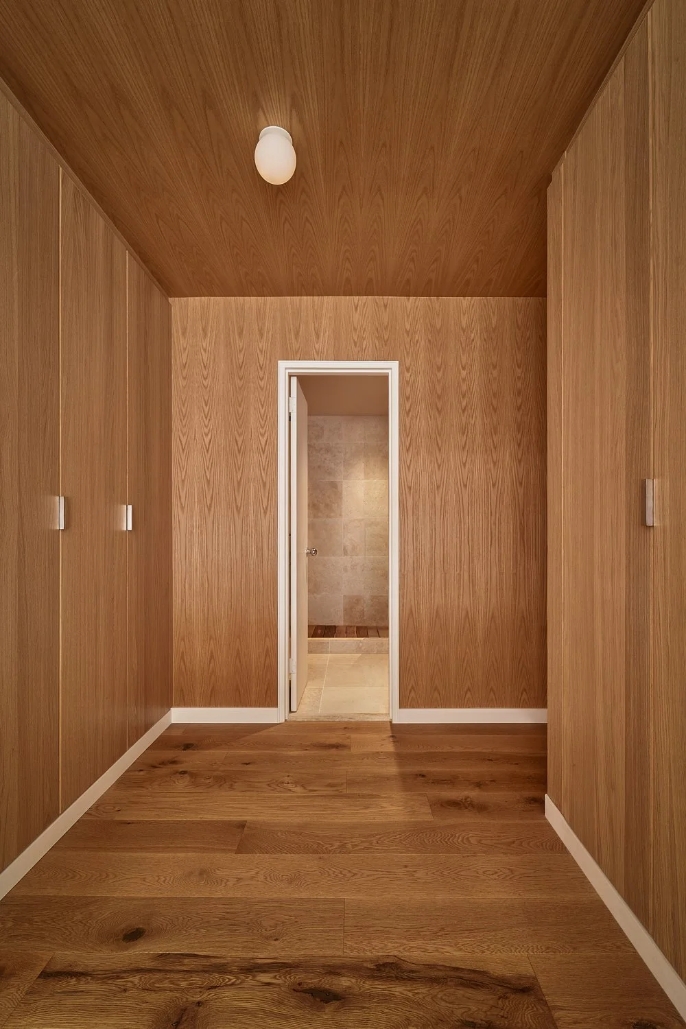 A narrow hallway with wood-paneled walls, ceiling, and floor, leading to a door that opens to a tiled bathroom.