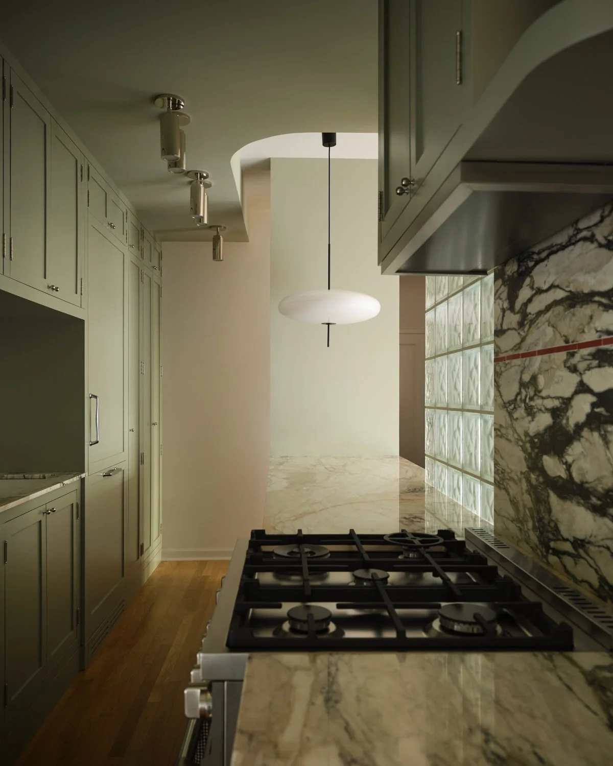 Modern kitchen with marble countertops, a gas stove, green cabinets, a hanging light fixture, and a glass block wall.